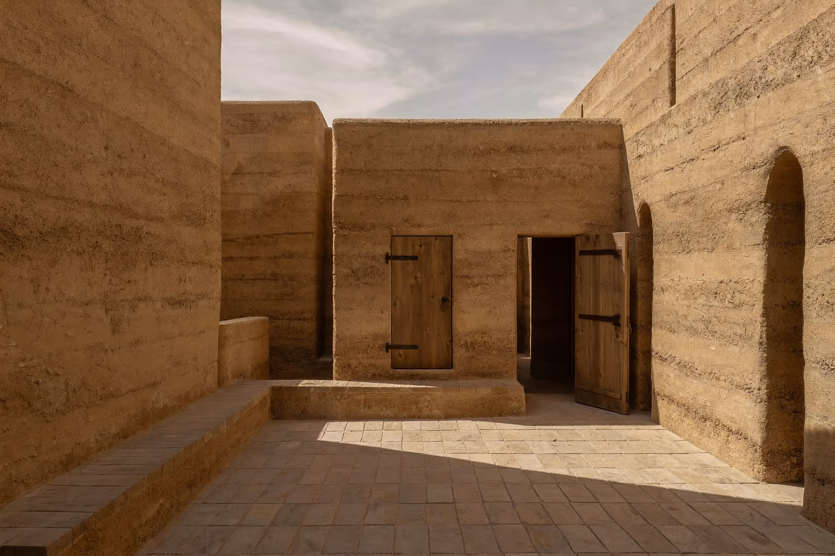 Cour intérieure encadrée de hauts murs en pisé aux strates horizontales visibles, sol en briques, deux portes en bois brut avec ferrures sombres, une arche creusée dans le mur de droite, lumière oblique de fin de journée, ciel légèrement voilé.