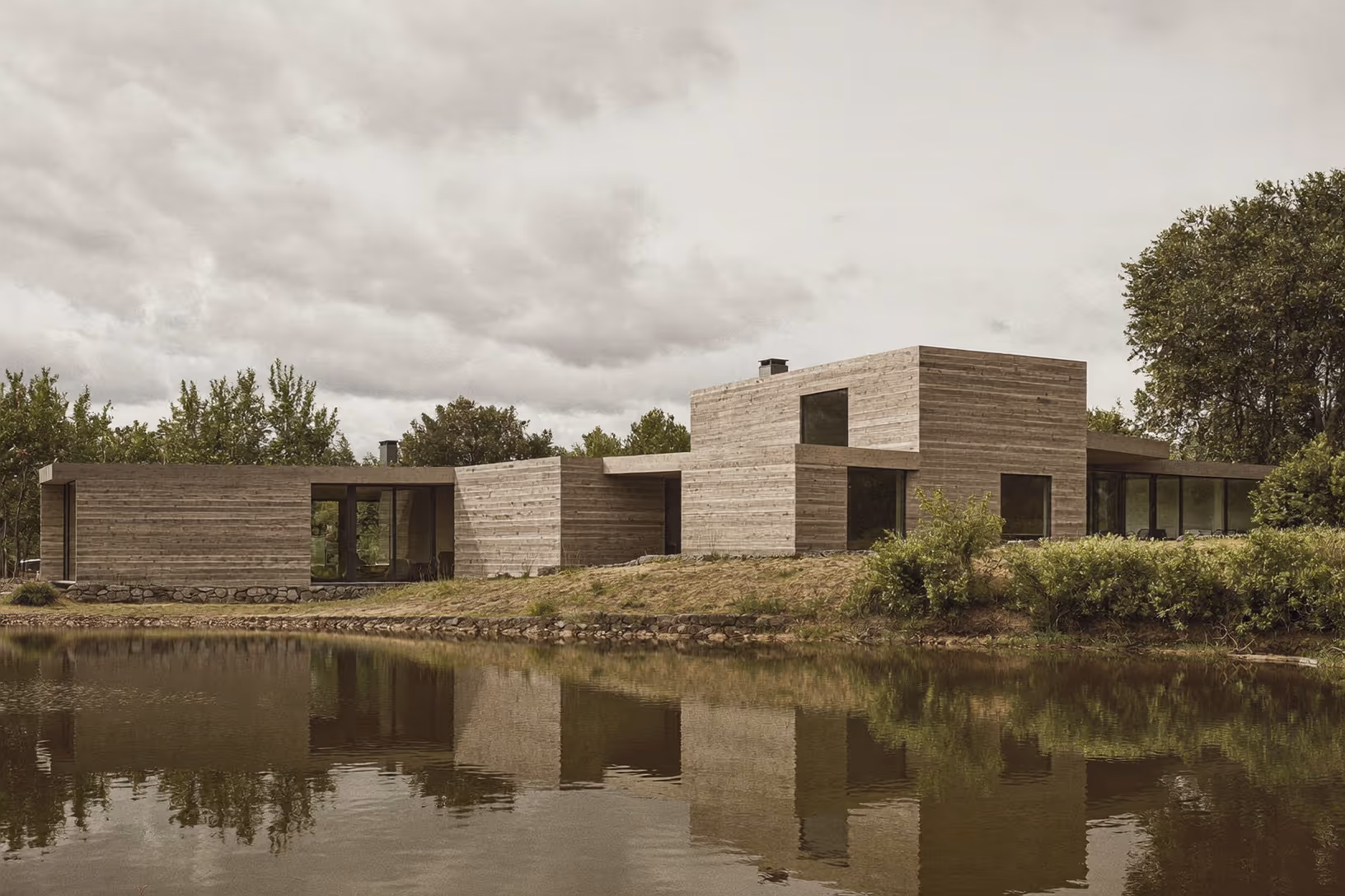 Maison composée de volumes cubiques horizontaux en bardage bois grisé, reflétée dans un plan d'eau calme, entourée d'arbres feuillus sous un ciel nuageux.