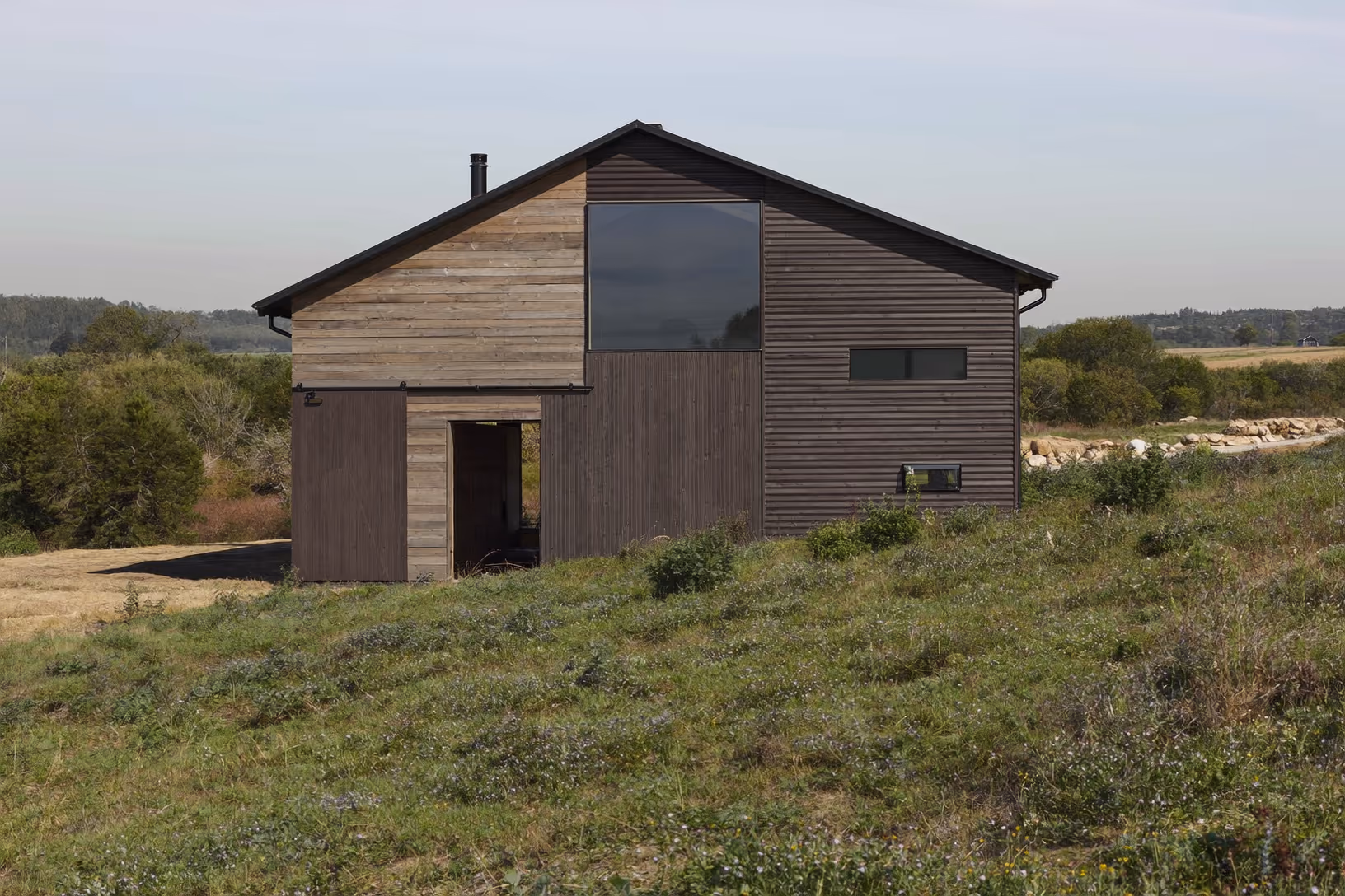 Maison à deux pans de toit recouverte d'un bardage horizontal en bois vieilli brun, grande baie vitrée réfléchissante en pignon, conduit de poêle en métal noir, posée dans une prairie de végétation basse avec une ligne d'arbres en arrière-plan.