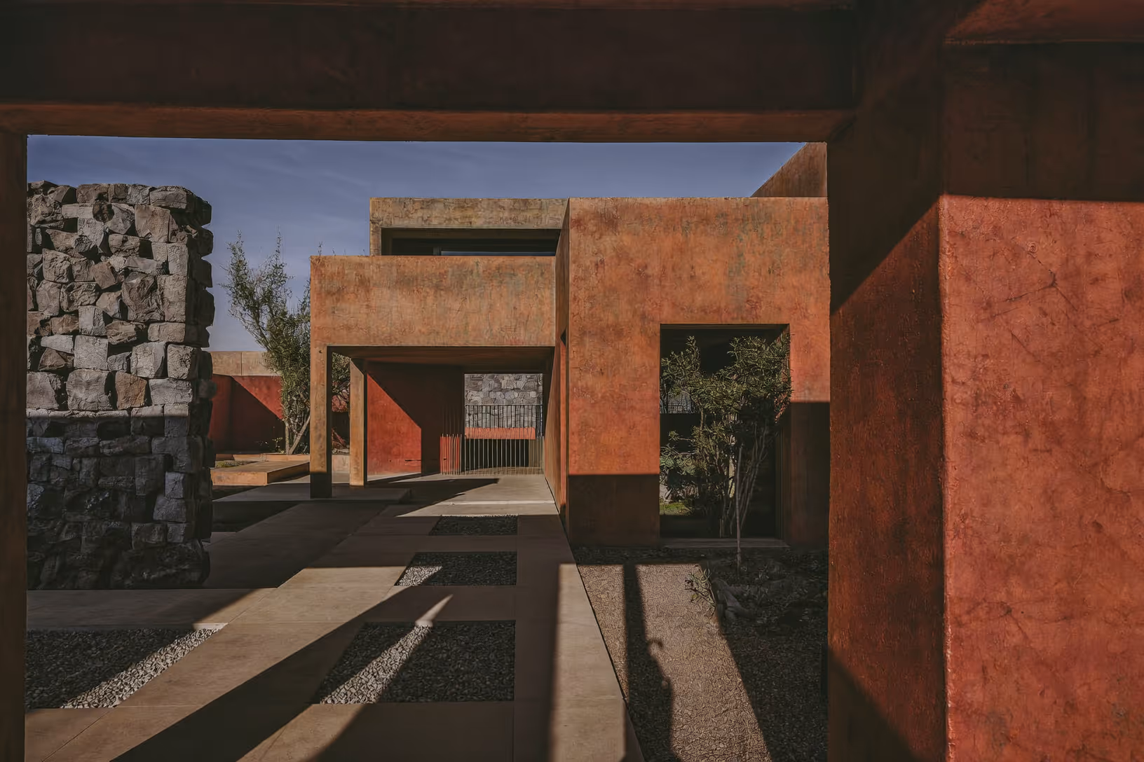 Cour intérieure encadrée par des volumes en béton teinté rouge-ocre, ombres portées sur le sol en béton, oliviers et graviers noirs, mur de pierres volcaniques brutes à gauche, ouvertures rectilignes en enfilade.