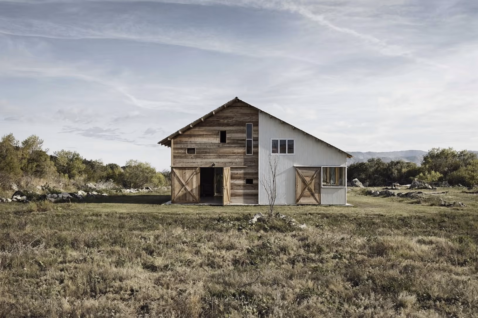 Façade d'une grange transformée, partagée entre bardage de bois de récupération patiné et revêtement métallique ondulé gris pâle, avec grandes portes coulissantes en croix, posée dans une prairie ouverte face à des collines boisées.