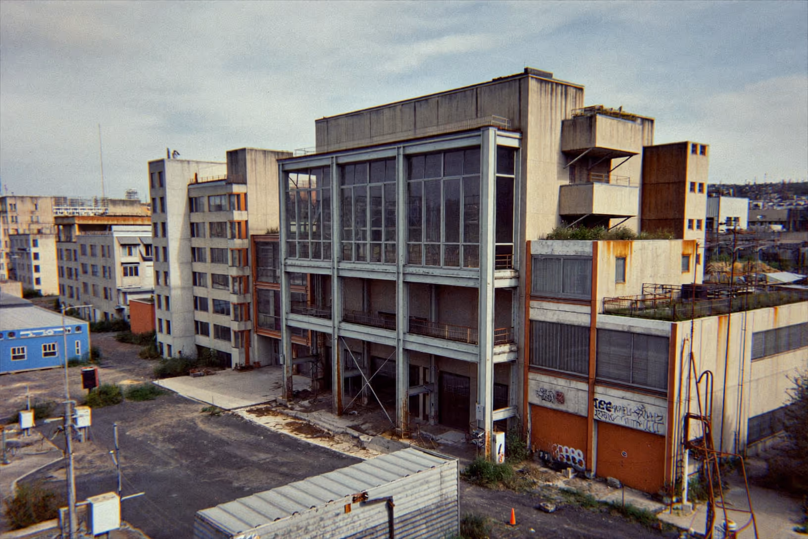 Vue en légère plongée d'un complexe de bâtiments industriels abandonnés, ossature béton brut, façade centrale vitrée sur structure métallique peinte en bleu-gris, parements en tôle rouillée, graffitis au niveau du sol, végétation spontanée sur les toits-terrasses, ciel couvert.