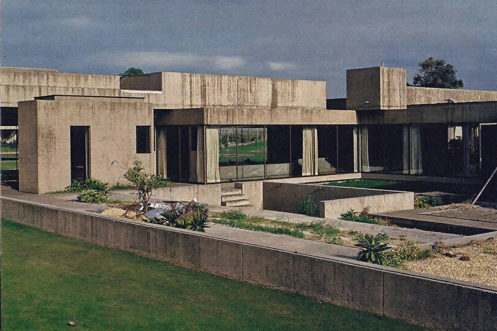 Façade d'une maison basse en béton brut apparent, vue depuis une pelouse verte, avec des volumes cubiques alternant pleins et vides, un jardin en creux au centre, de larges baies vitrées à rideaux blancs, et un ciel couvert aux nuages sombres.