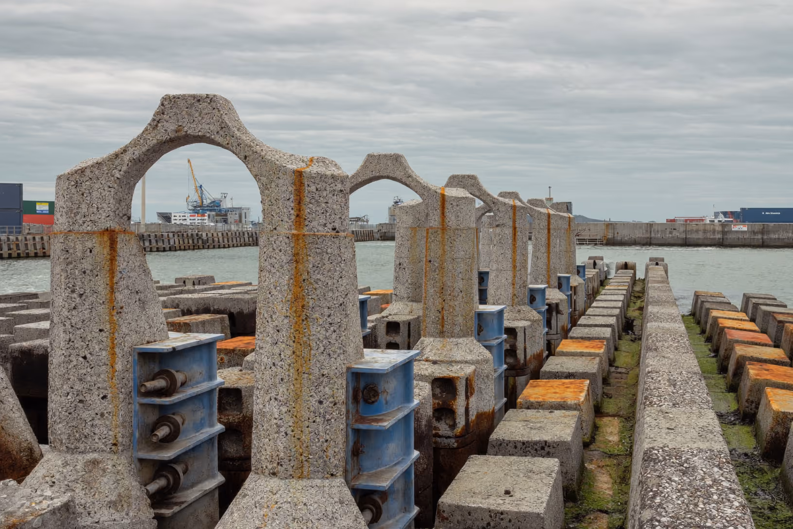 Rangée de piliers en béton à arches évidées sur un quai portuaire, reliés par des connecteurs métalliques bleus et rouillés, avec la mer et des grues industrielles en arrière-plan sous un ciel couvert.
