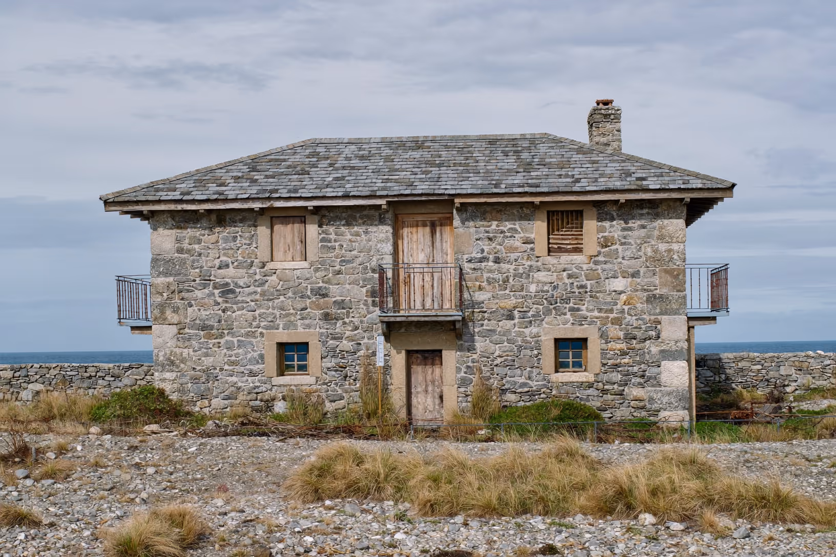 Maison de deux étages en pierres grises appareillées, toit en ardoise à quatre pans, volets et porte en bois vieilli, deux balcons en fer forgé, petites fenêtres à petits-bois bleus, sol caillouteux aride au premier plan, horizon marin sous ciel couvert.