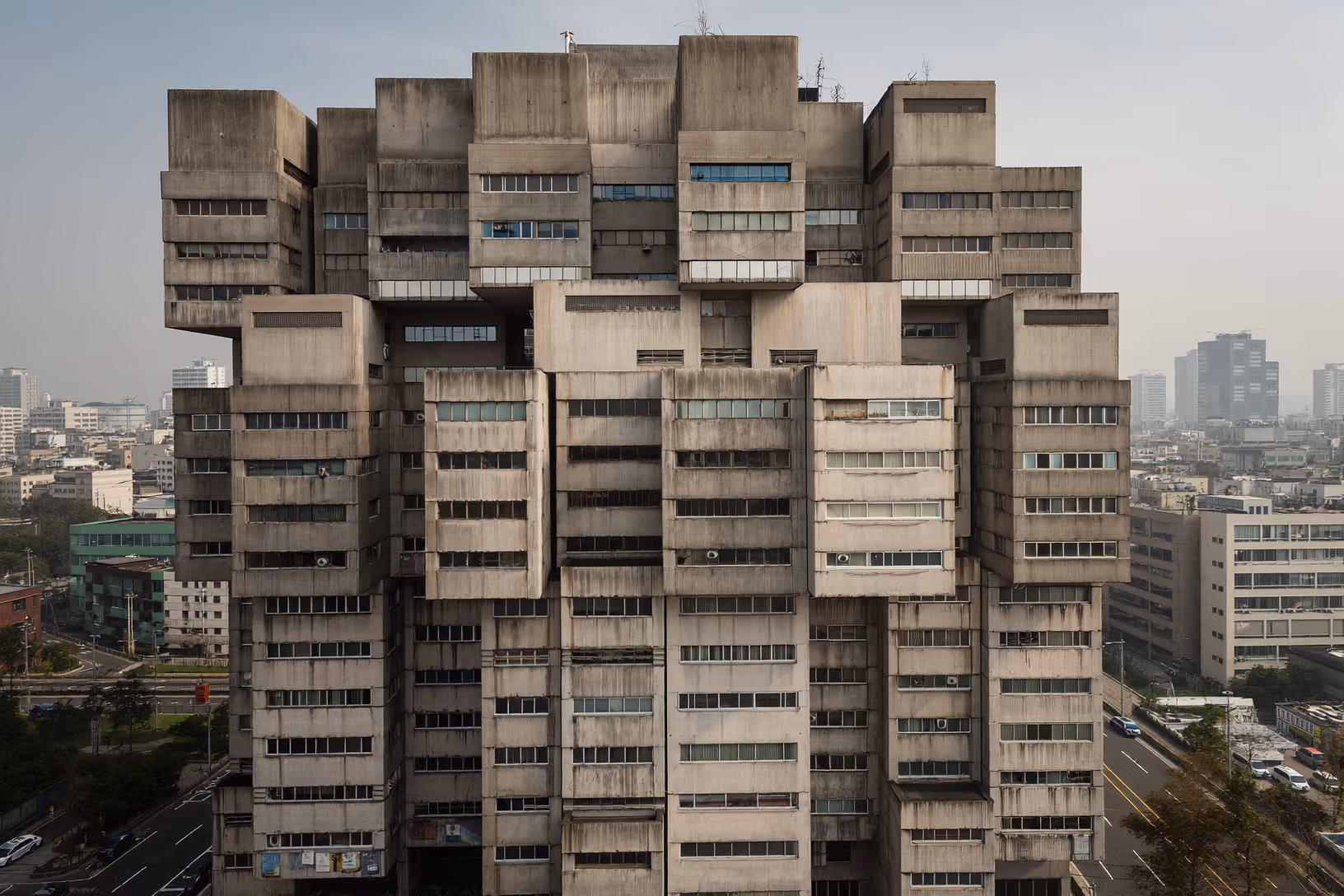 Tour brutaliste en béton apparent composée de volumes cubiques décalés et superposés, photographiée en vue aérienne légèrement plongeante sur fond de ville dense sous ciel voilé.