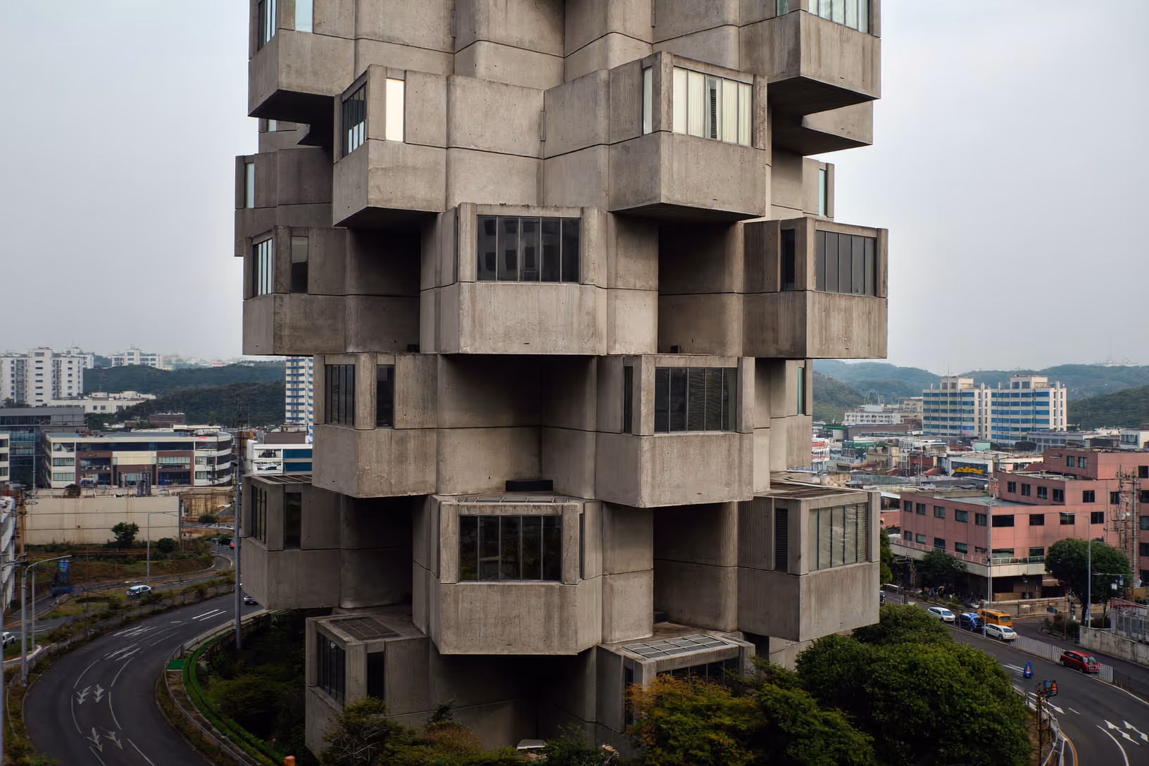 Tour de béton brut aux volumes modulaires en saillie, photographiée en contre-plongée légère, sur fond de ciel gris et de ville périphérique.