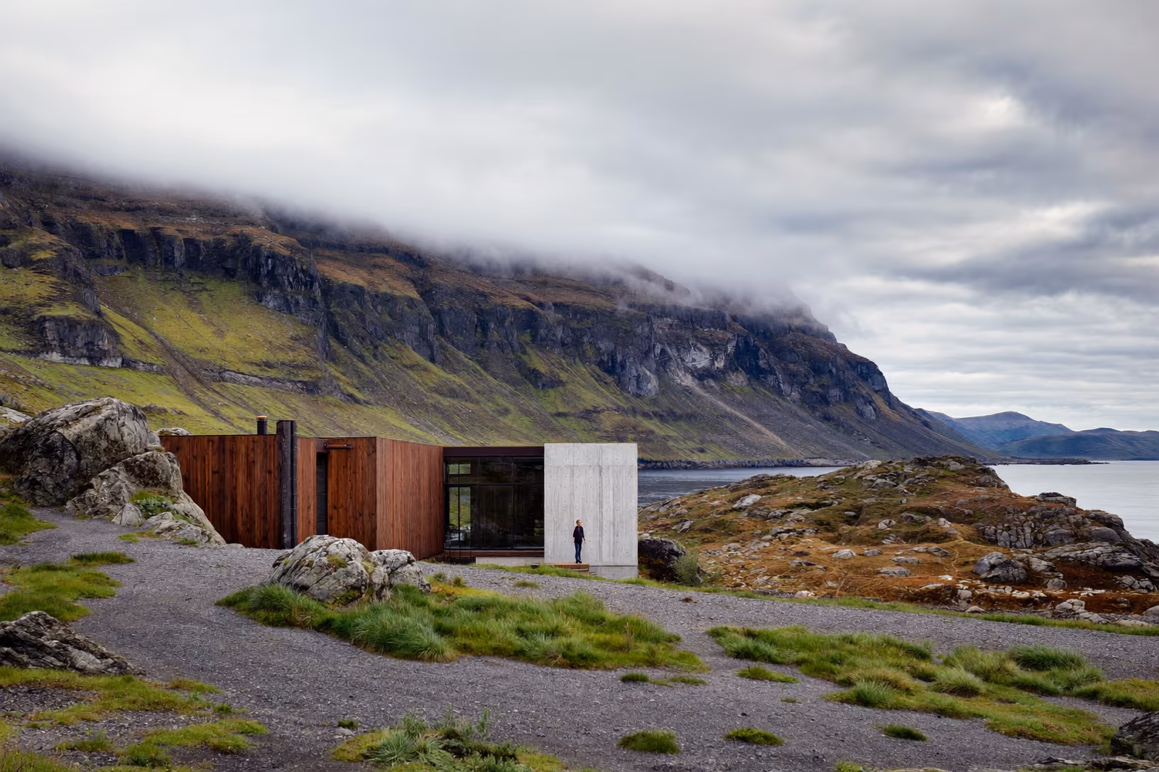 Construction basse à ossature de bois sombre et volume en béton brut, posée sur un sol rocheux face à un fjord, au pied de falaises verdoyantes enveloppées de nuages, une silhouette humaine debout devant la façade.