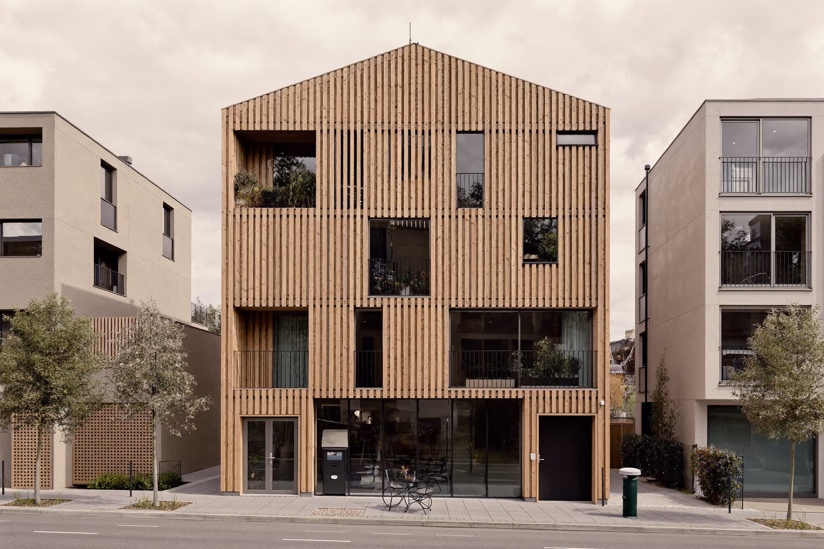 Façade frontale d'un bâtiment urbain de trois niveaux entièrement recouvert de lames verticales en bois clair, toit en pignon, encadré par deux immeubles en béton enduit, sur une rue de ville européenne sous ciel couvert.
