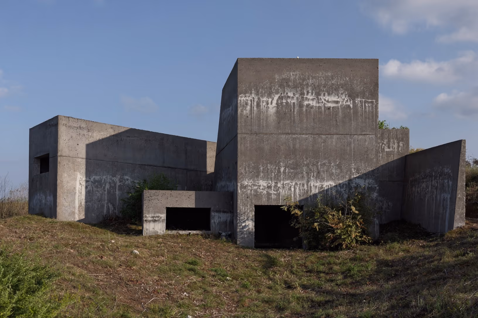 Ensemble de volumes cubiques en béton brut sur une colline herbeuse, avec de rares ouvertures basses et des traces d'efflorescence sur les parois, ciel bleu en arrière-plan.