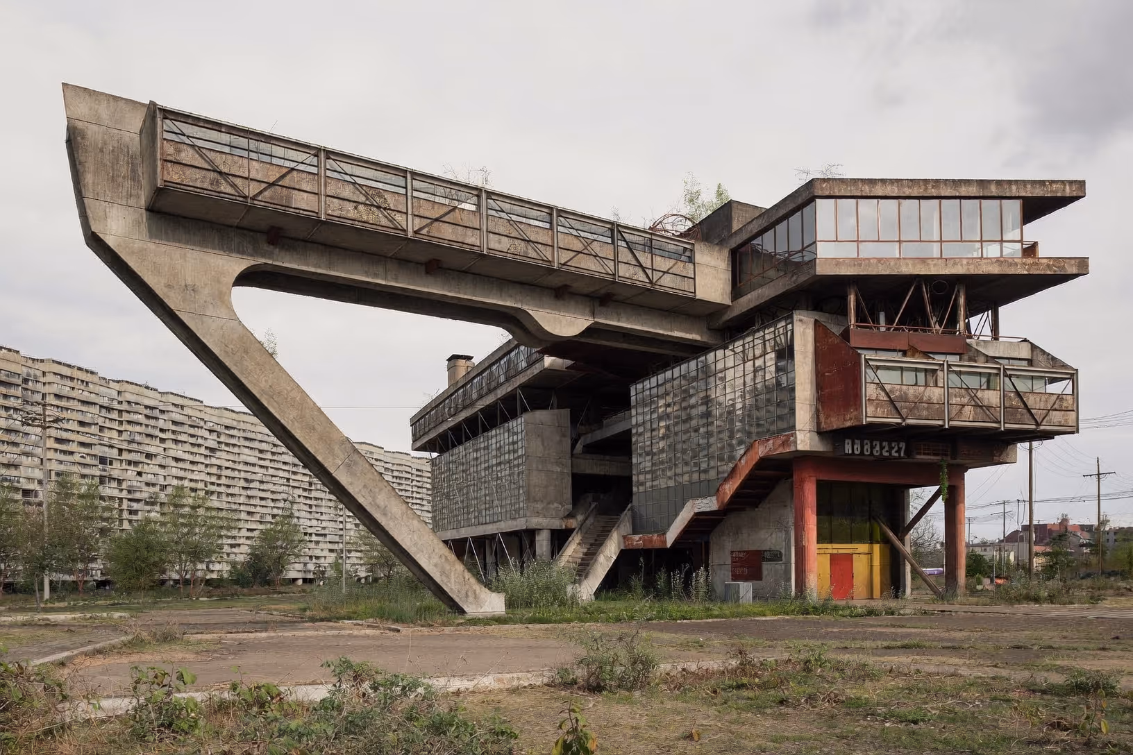 Bâtiment brutaliste abandonné avec passerelle en béton portée par un pilier en V, volumes vitrés oxydés et barres d'immeubles en arrière-plan sous ciel nuageux.