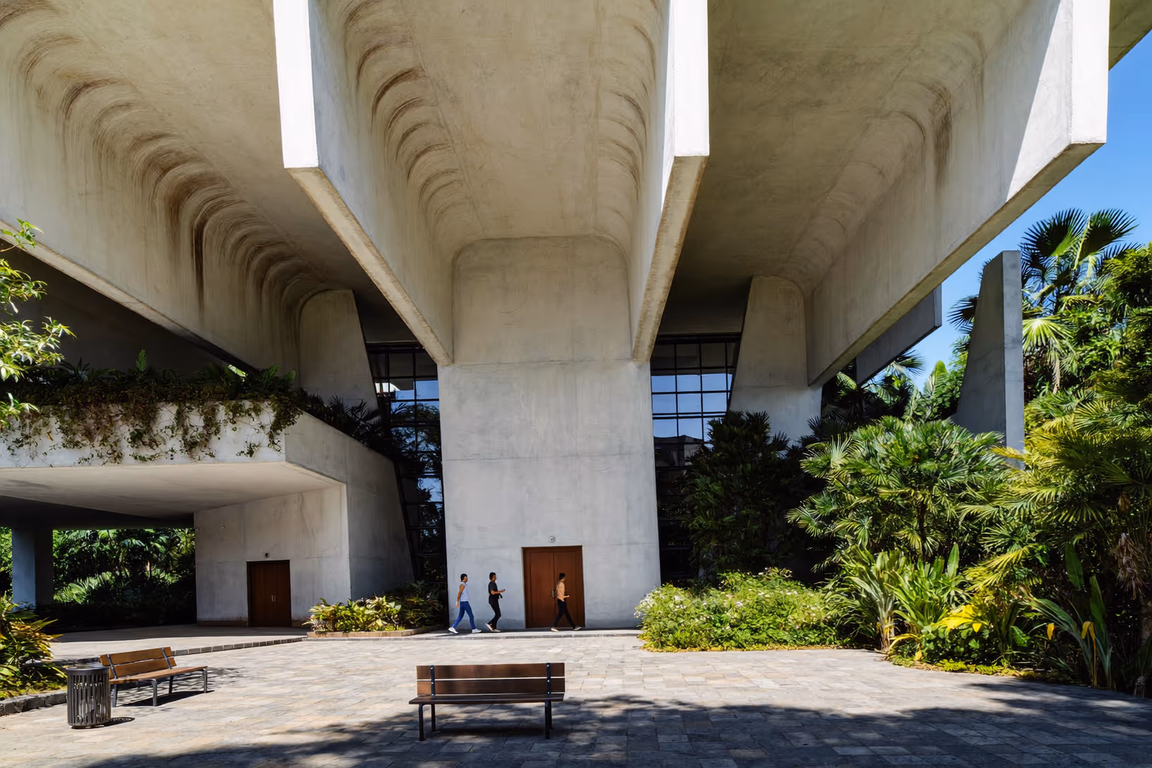 Sous-face nervurée d'un bâtiment brutaliste en béton brut, soulevé sur de larges piliers sculptés ; végétation tropicale dense en arrière-plan, deux silhouettes humaines au centre.