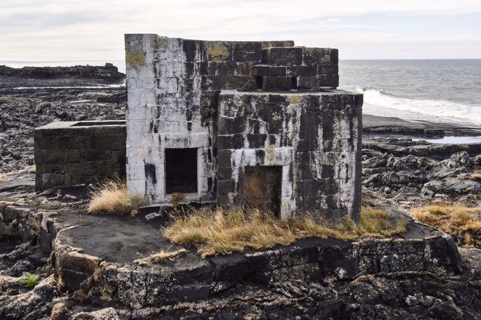 Structure en blocs de pierre taillée sombre, patinée de lichens blancs, posée sur un socle rocheux côtier. Herbes sèches au pied des murs. Mer agitée et ciel couvert en arrière-plan.