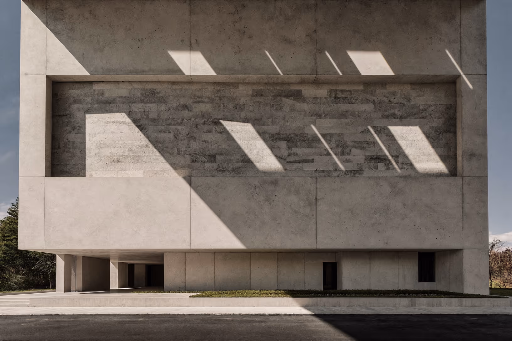 Façade d'un grand volume en béton banché surélevé sur piliers, avec une bande centrale en pierre appareillée grise ; des ombres obliques et parallèles projetées par le soleil rasant couvrent la surface du mur.