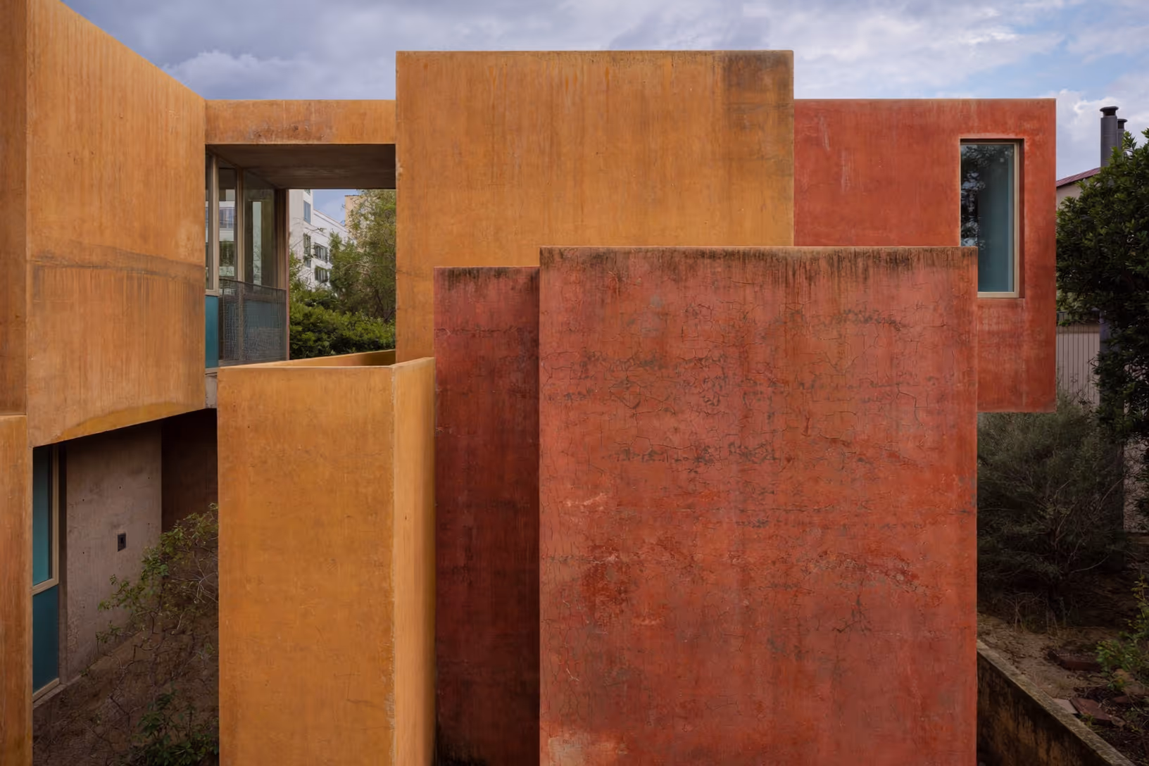 Deux volumes de béton enduit orthogonaux, l'un ocre doré et l'autre terracotta, se superposent en décalé. De rares ouvertures verticales encadrées de turquoise percent les parois. Végétation et ciel nuageux apparaissent dans les interstices.