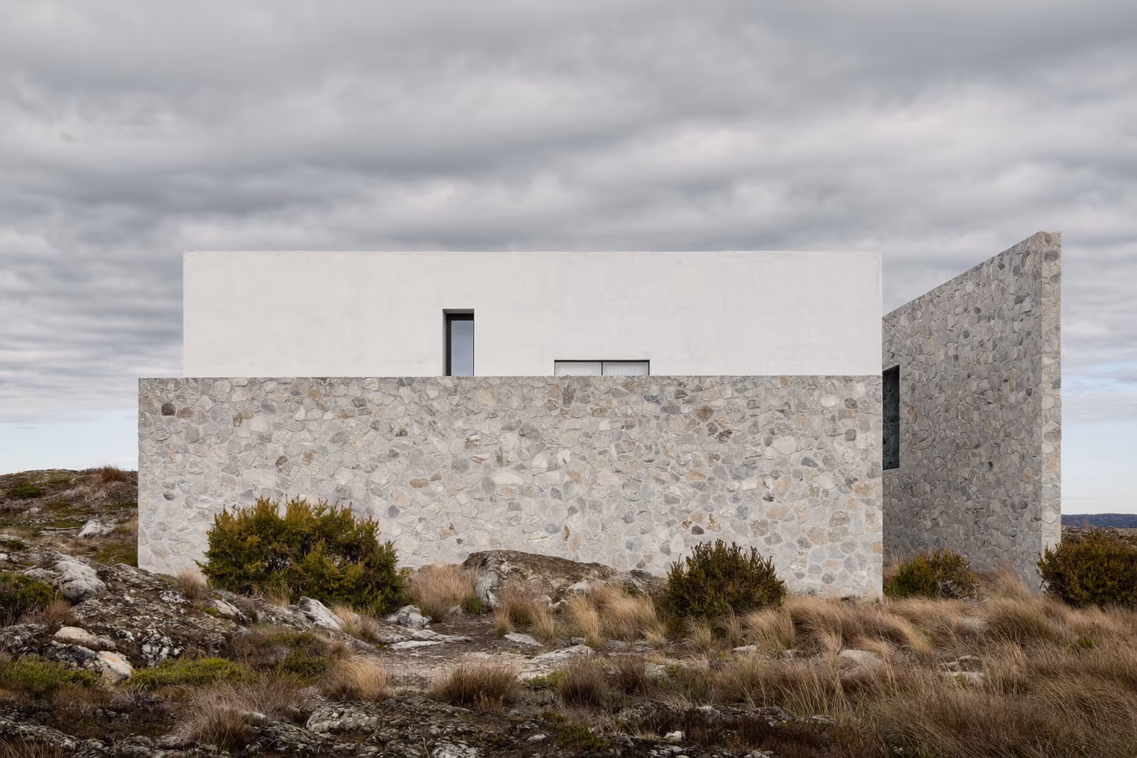 Façade d'une maison sur plateau rocheux : mur bas en galets apparents, volume supérieur enduit blanc mat, contrefort de pierre à droite, ciel couvert, végétation rase.