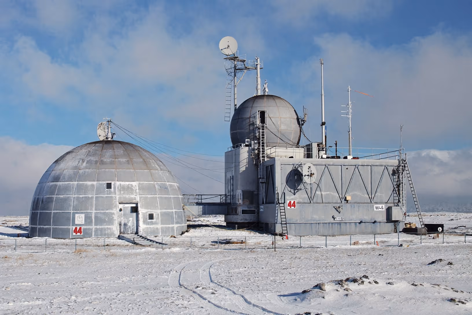 Deux structures géodésiques en métal gris, équipées d'antennes et de mâts de communication, sur un sol enneigé et plat sous un ciel nuageux.