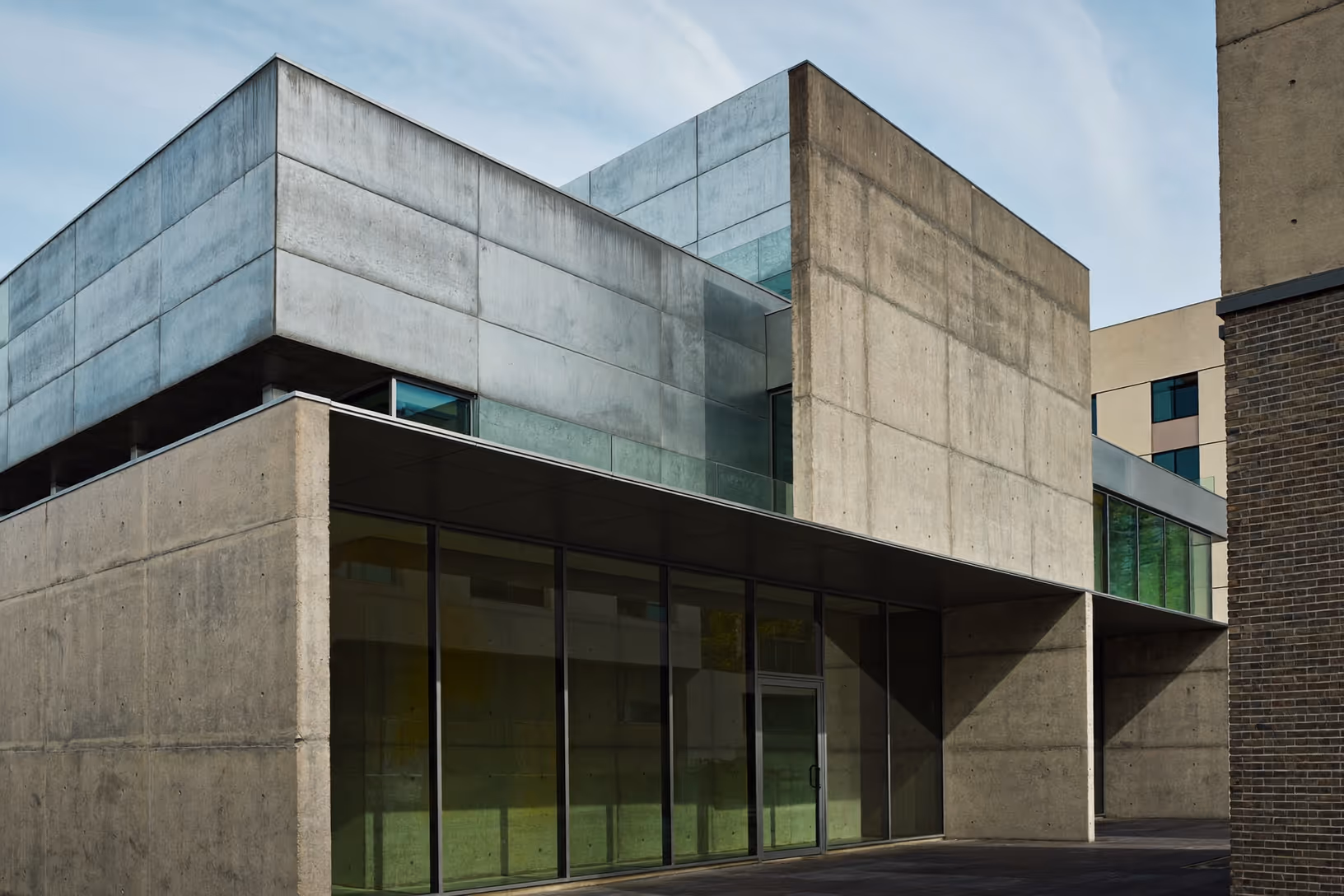 Bâtiment en béton brut à deux niveaux : un volume supérieur en porte-à-faux repose sur un socle entièrement vitré à menuiseries noires, vitrage teinté vert reflétant la végétation, lumière rasante de fin d'après-midi, ciel bleu dégagé.