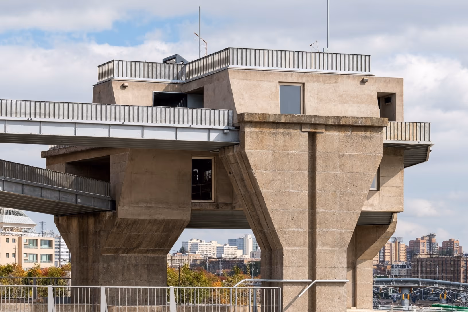 Structure brutaliste en béton brut élevée sur de larges piliers trapézoïdaux, traversée par deux passerelles métalliques horizontales, sur fond de skyline urbaine et ciel nuageux.