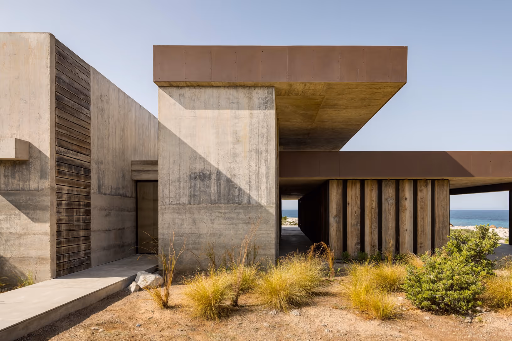 Façade en béton banché et lattes de bois grisé, avec deux toits-dalles en porte-à-faux recouverts d'acier corten rouille, des colonnes en bois debout sous un auvent ouvert côté mer ; au premier plan, touffes de graminées sèches et arbustes bas sur sol sableux aride.