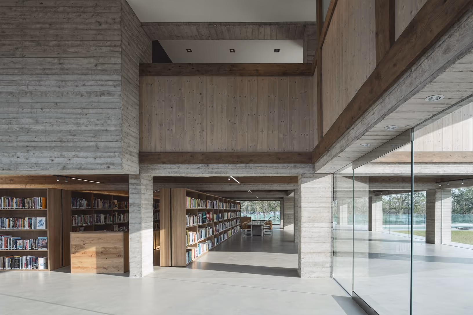 Intérieur d'une bibliothèque à double hauteur en béton brut coffré au bois, avec des rayonnages en chêne clair, un sol en béton poli et une paroi vitrée ouvrant sur un jardin arboré.