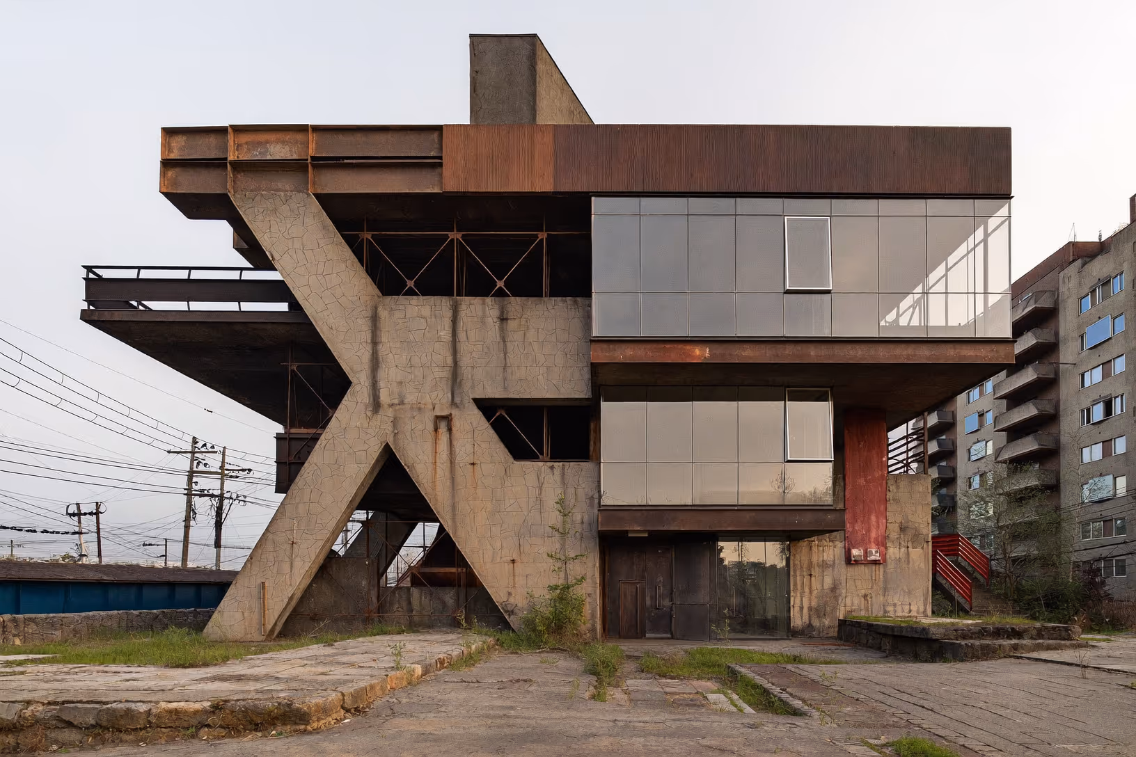 Bâtiment abandonné à structure brutaliste : un gigantesque contrefort en X de béton brut soutient un volume supérieur habillé d'acier Corten et d'un bandeau vitré. Escaliers métalliques rouges à droite, sol en asphalte fissuré, immeuble résidentiel en arrière-plan, ciel gris.