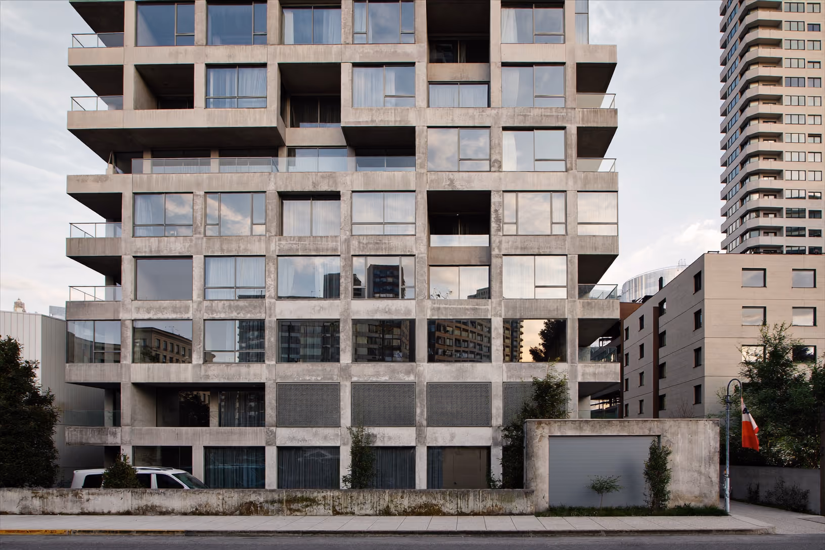 Façade d'une tour de béton gris en milieu urbain, organisée en trame orthogonale de cadres structurels et de baies vitrées, avec quelques balcons et panneaux opaques, végétation basse au sol, ciel couvert.