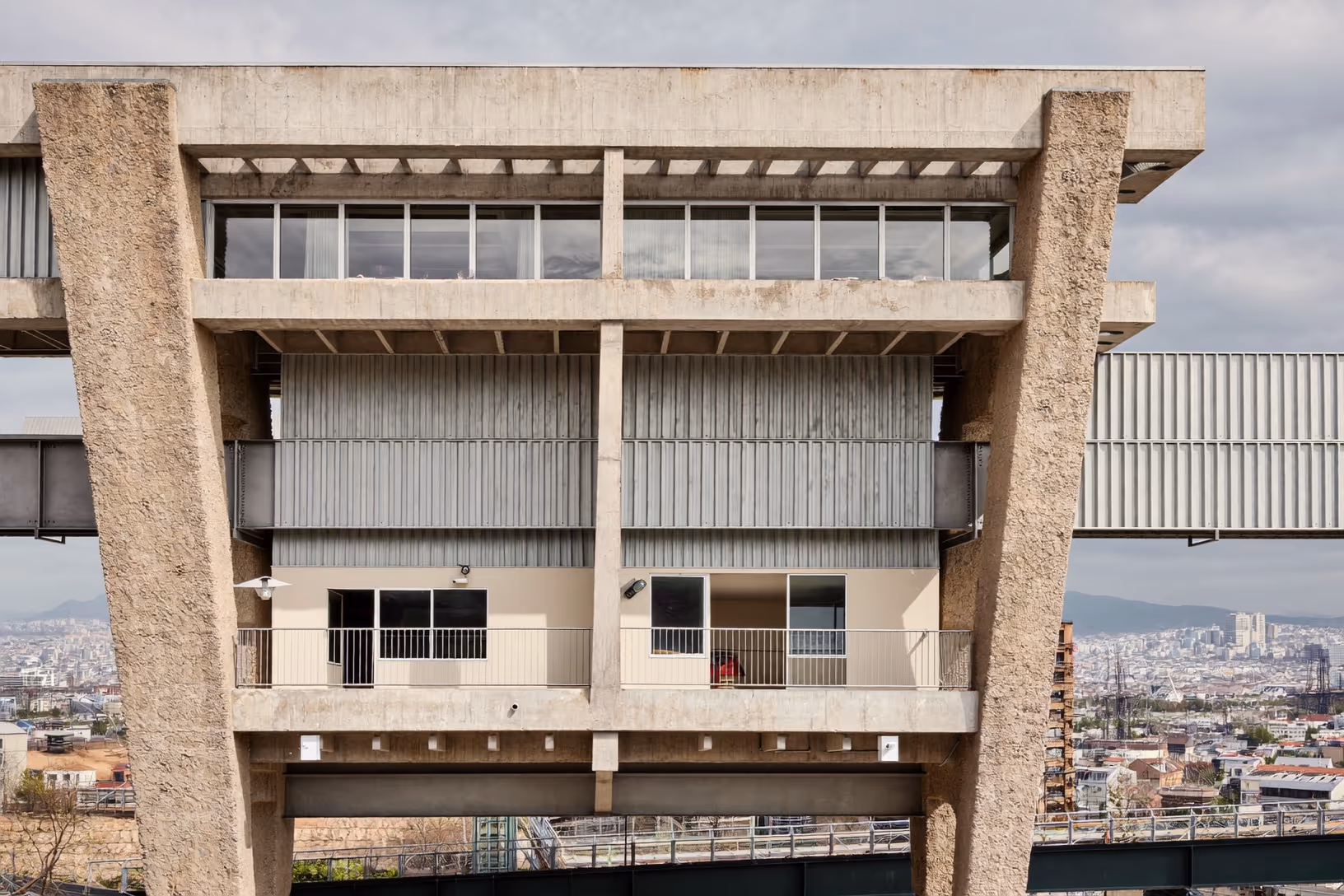 Façade brutaliste en béton brut vue de face, composée de deux piliers massifs encadrant trois niveaux superposés — vitrage continu en bandeau au sommet, bardage métallique strié au centre, balcons à garde-corps fins en bas — avec une ville dense en arrière-plan sous un ciel couvert.