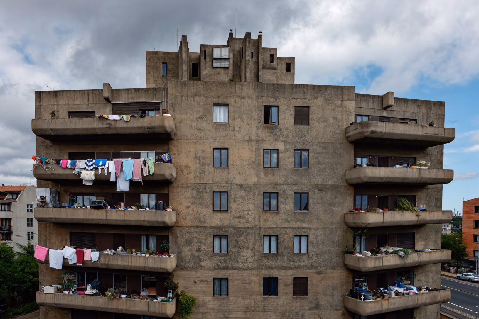 Façade frontale d'un immeuble collectif en béton brut à sept niveaux, avec balcons arrondis en porte-à-faux de part et d'autre, linge coloré étendu, plantes en pot sur les garde-corps, fenêtres rectangulaires régulièrement distribuées, et une couronne de volumes découpés au sommet, sur fond de ciel nuageux.