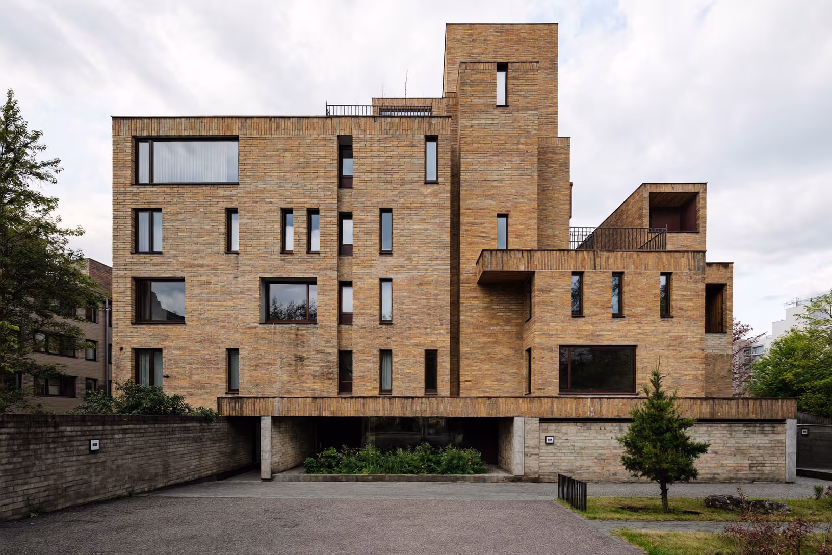Façade d'un immeuble en brique ocre à quatre niveaux, composée de volumes cubiques décalés et superposés, avec des fenêtres verticales et horizontales de formats variés, élevé sur pilotis au-dessus d'un parvis minéral, encadré d'arbres sous un ciel nuageux.