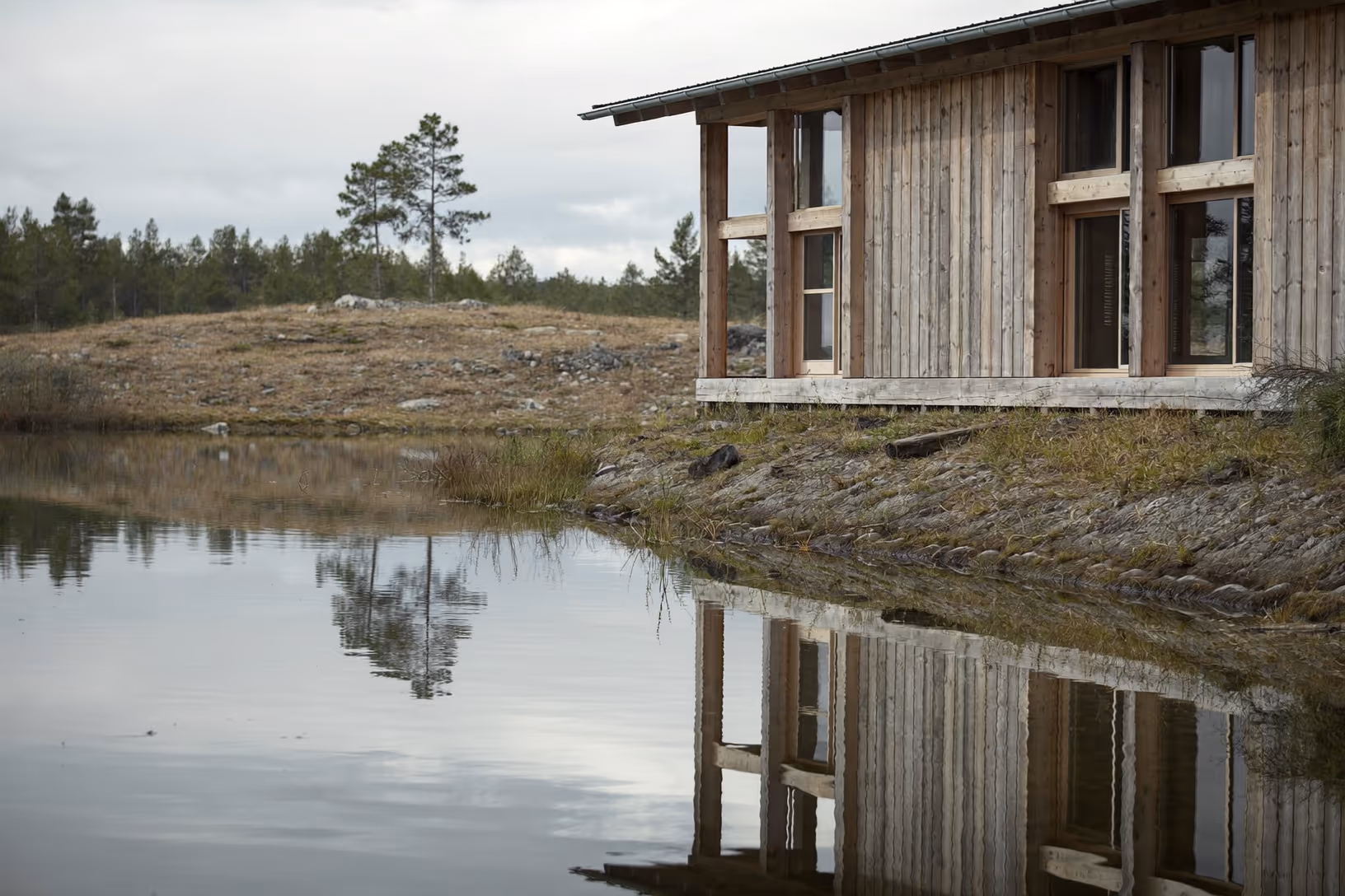 Pavillon en bardage bois vertical grisé au bord d'un lac calme, entouré d'une lande rocheuse et de pins épars sous un ciel couvert, avec le reflet de la façade dans l'eau immobile.