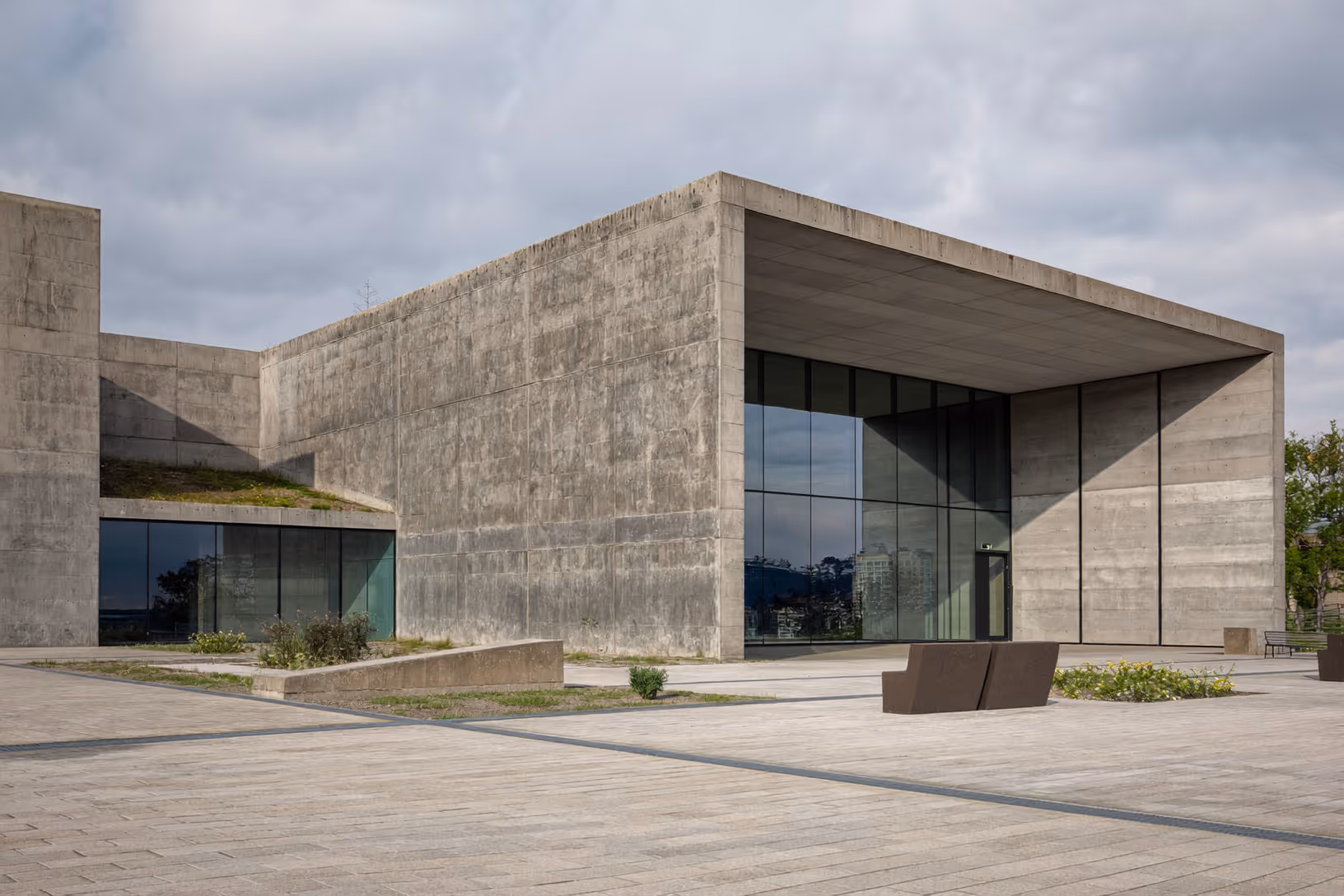 Édifice en béton brut apparent avec large porte-à-faux sur façade vitrée, toiture végétalisée sur volume bas à gauche, esplanade en dalles de pierre et bancs en acier corten au premier plan, ciel nuageux.