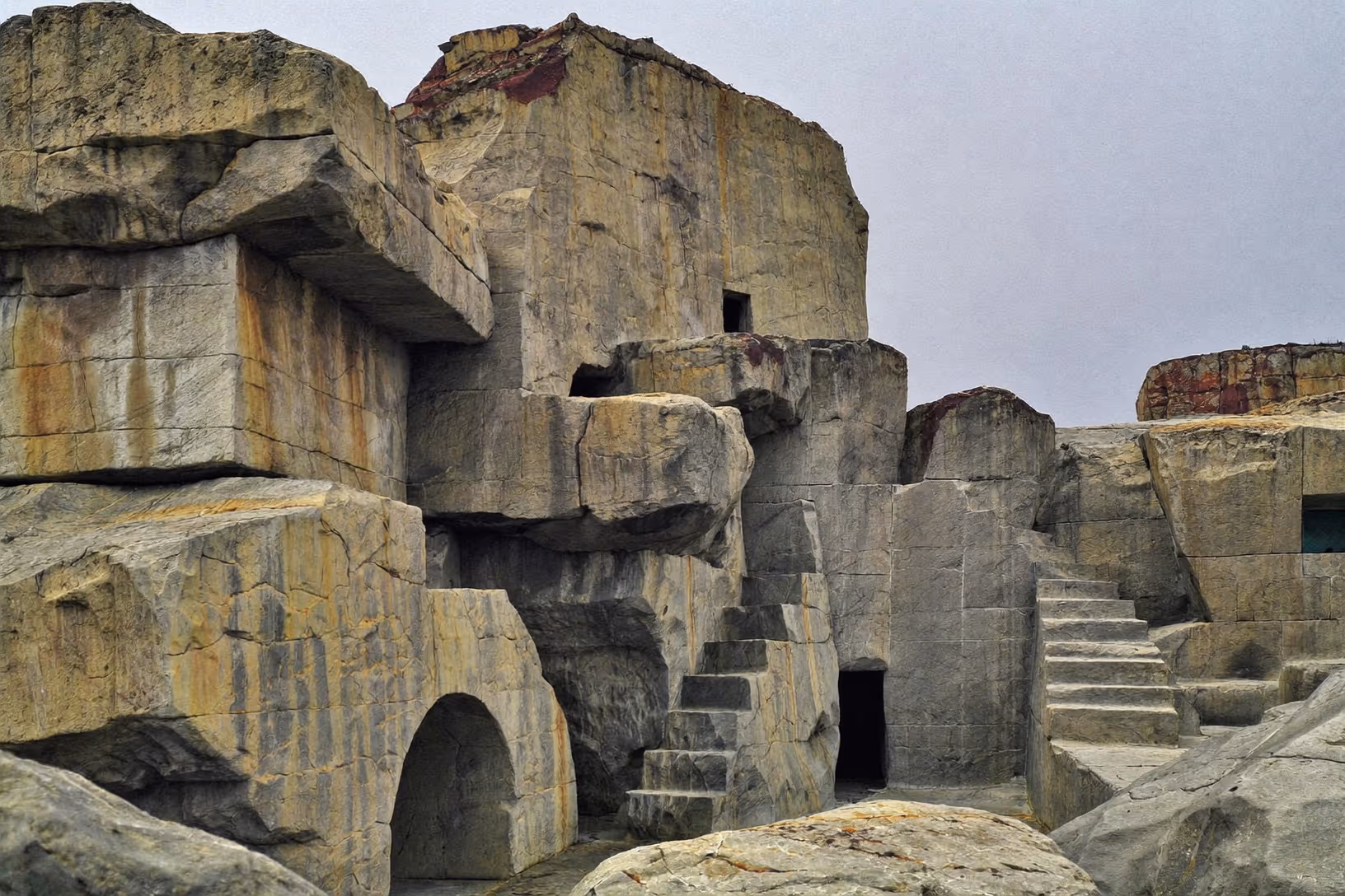 Structure monumentale taillée dans des blocs de roche grise et ocre, avec escaliers creusés, ouvertures en arc et fenêtres étroites, sous un ciel couvert uniforme.