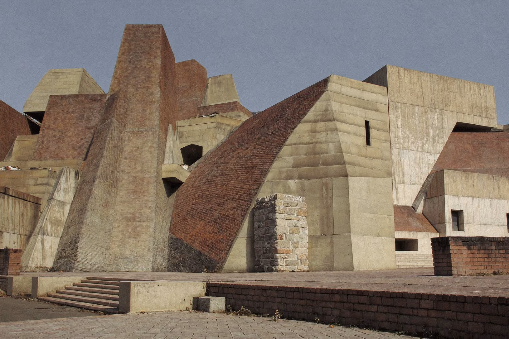 Ensemble architectural monumental composé de masses en béton brut et de parements en brique rouge — pyramides, arches inversées, volumes tronqués — sous un ciel gris pâle, photographié depuis une esplanade pavée déserte.