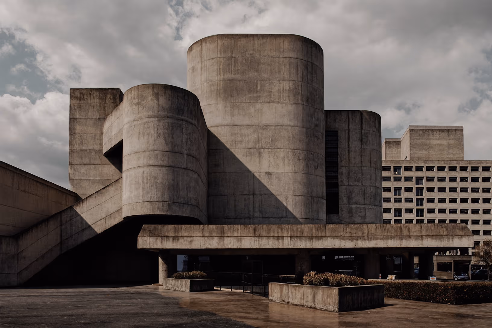 Édifice brutaliste en béton brut composé de plusieurs grands cylindres superposés sur un socle horizontal surélevé, photographié sous un ciel nuageux dramatique, avec un bâtiment en grille rectangulaire visible en arrière-plan.