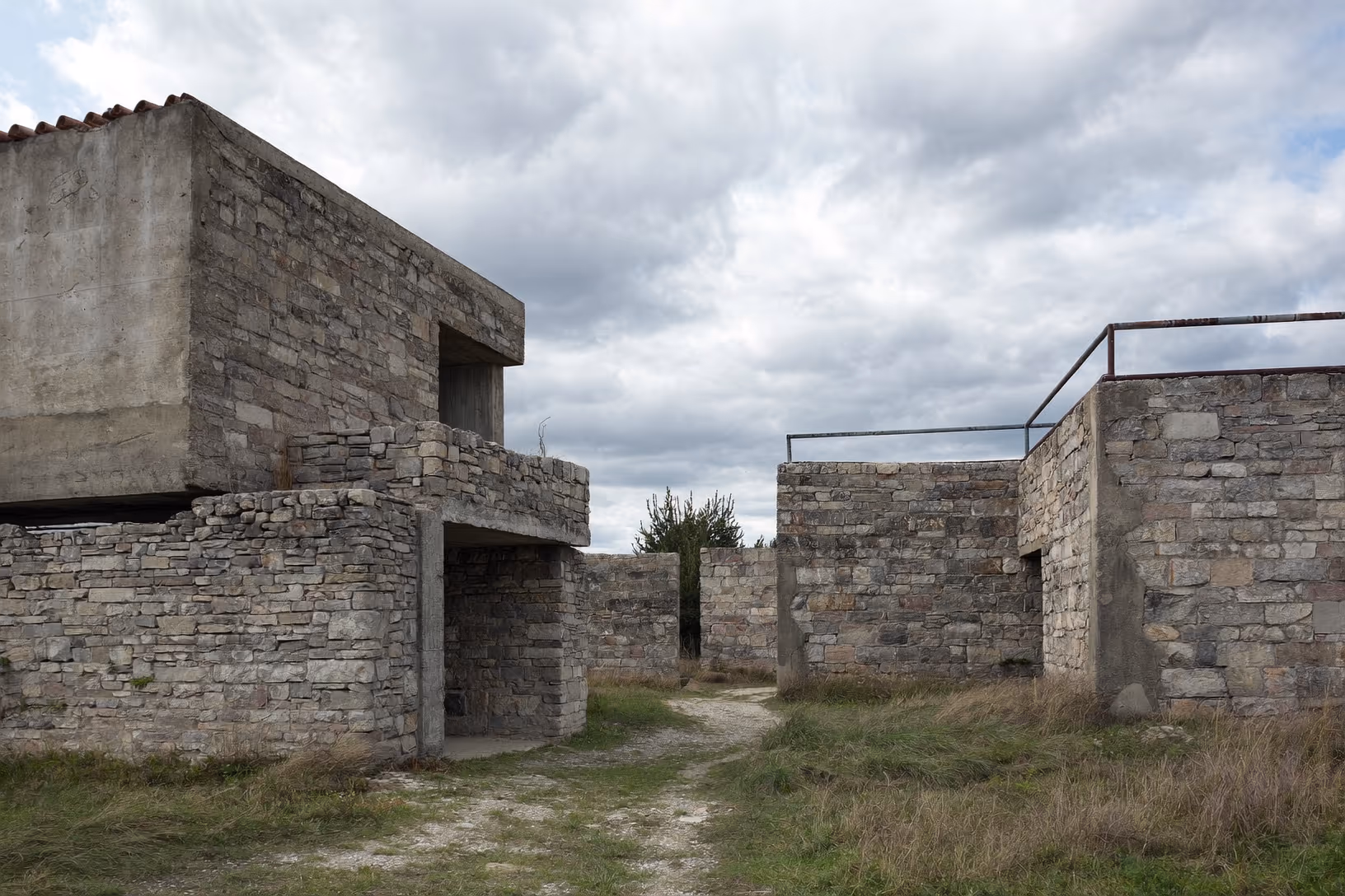 Deux volumes en pierre de taille grise reliés par un mur bas, avec un volume supérieur en béton brut et des ouvertures découpées dans la masse ; végétation sèche au sol, ciel nuageux chargé.