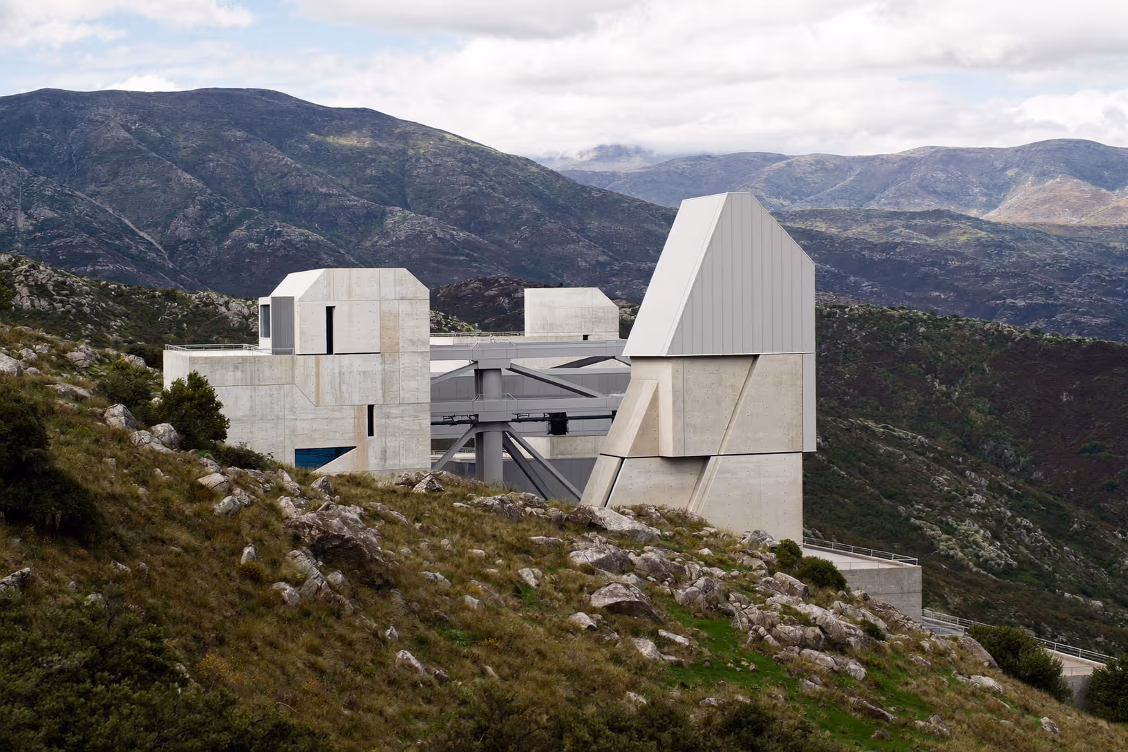 Édifice en béton brut composé de plusieurs volumes géométriques à facettes biais, reliés par une structure métallique inclinée, implanté sur une pente rocailleuse avec un arrière-plan de massif montagneux sous ciel couvert.