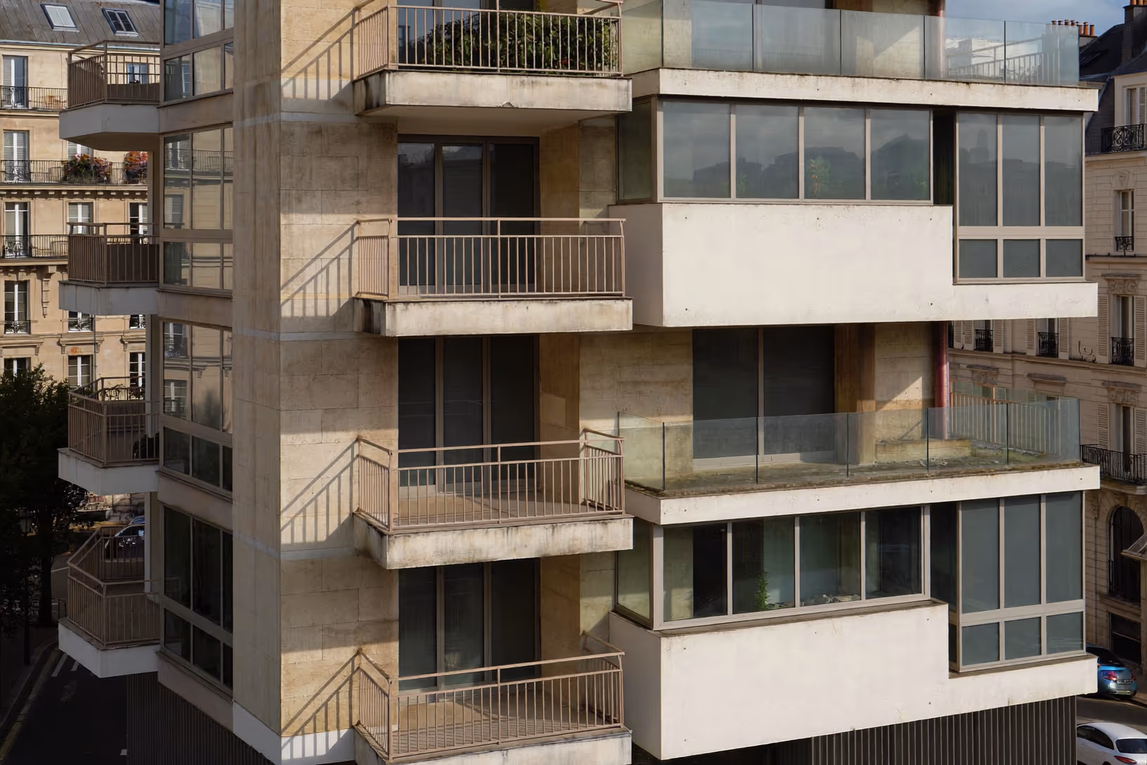 Façade d'un immeuble résidentiel contemporain en béton clair, avec balcons en porte-à-faux, garde-corps métalliques fins et larges baies vitrées, sur fond de tissu haussmannien.