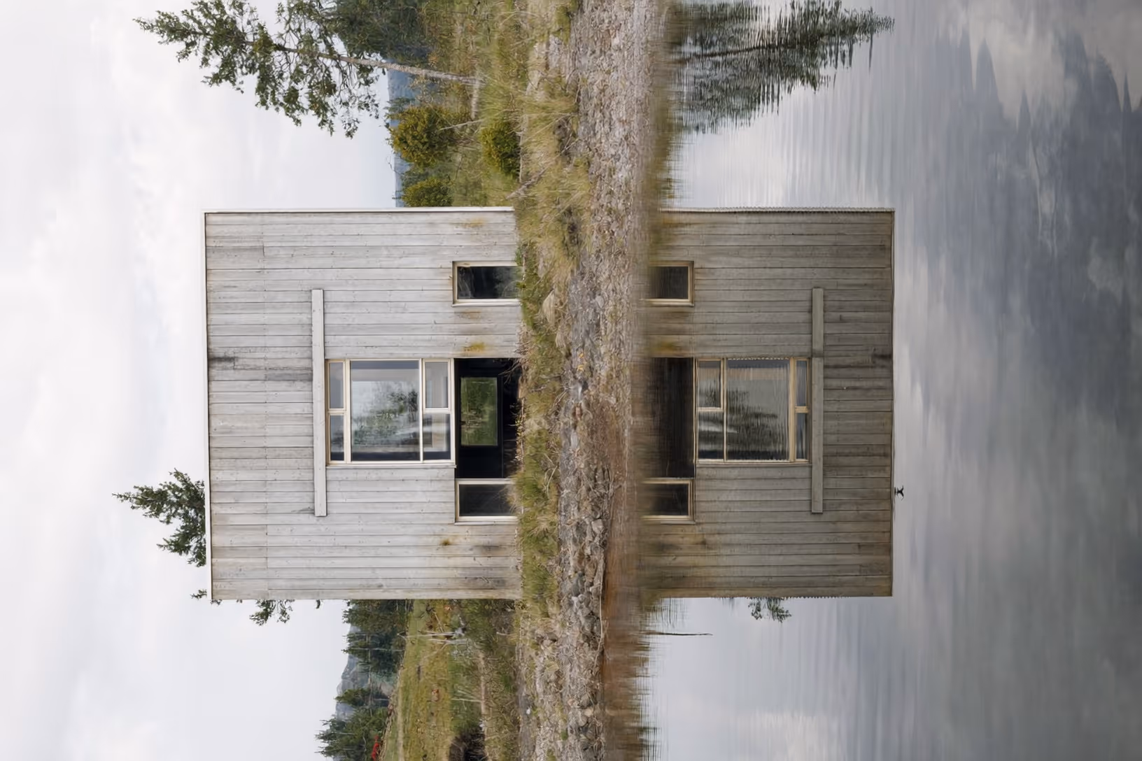 Façade frontale d'un petit volume rectangulaire en bardage bois horizontal grisé, posé sur une rive rocheuse et herbeuse au bord d'un lac calme sous un ciel couvert.