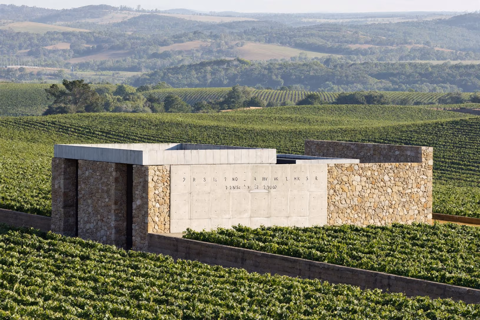 Bâtiment bas en pierre appareillée et béton brut, posé au milieu de vignes en rangées, collines boisées à l'arrière-plan sous une lumière diffuse.