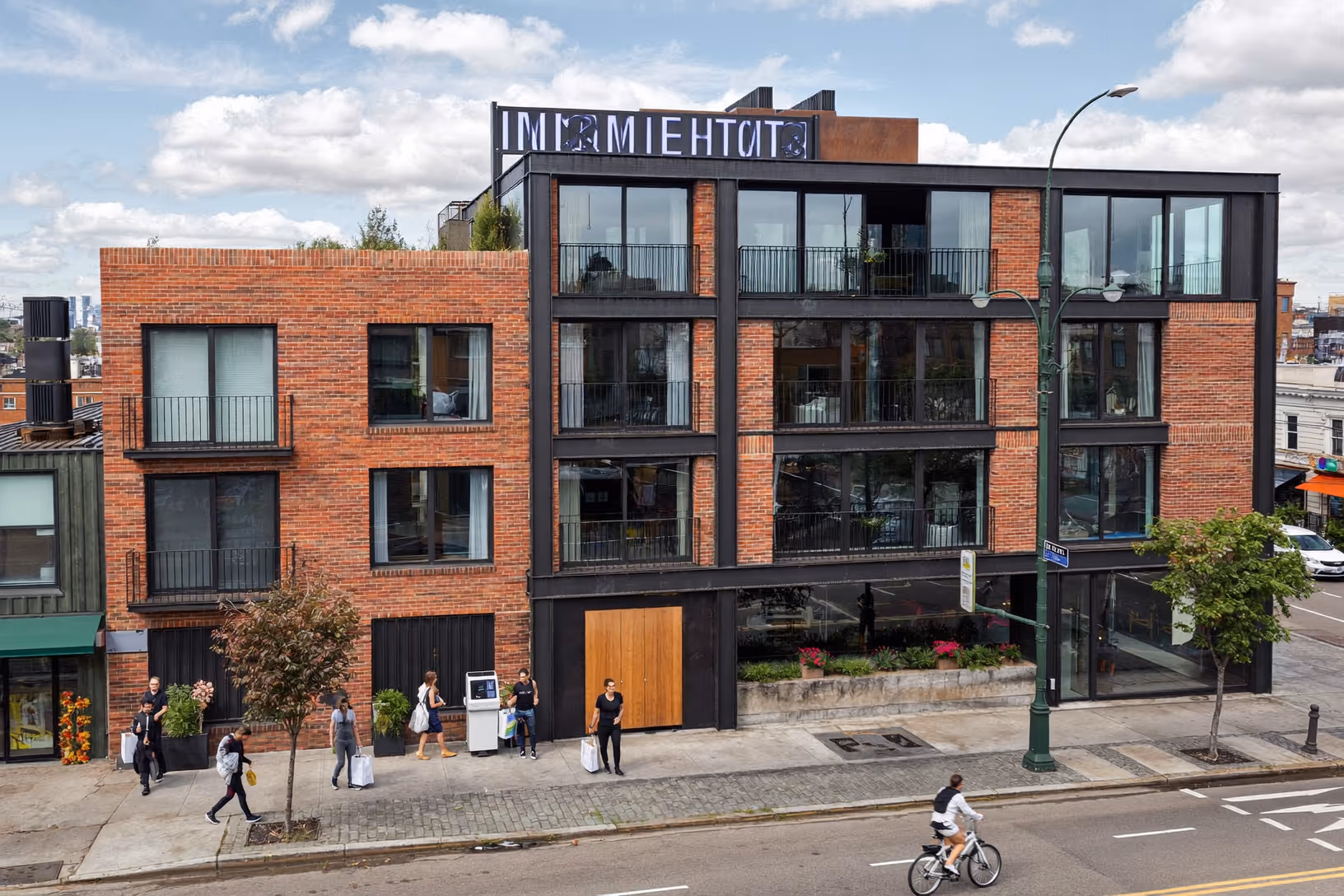 Immeuble de quatre niveaux en brique rouge et structure métallique noire, à l'angle d'une rue urbaine animée, avec de larges baies vitrées, une entrée en bois et une terrasse végétalisée en toiture.