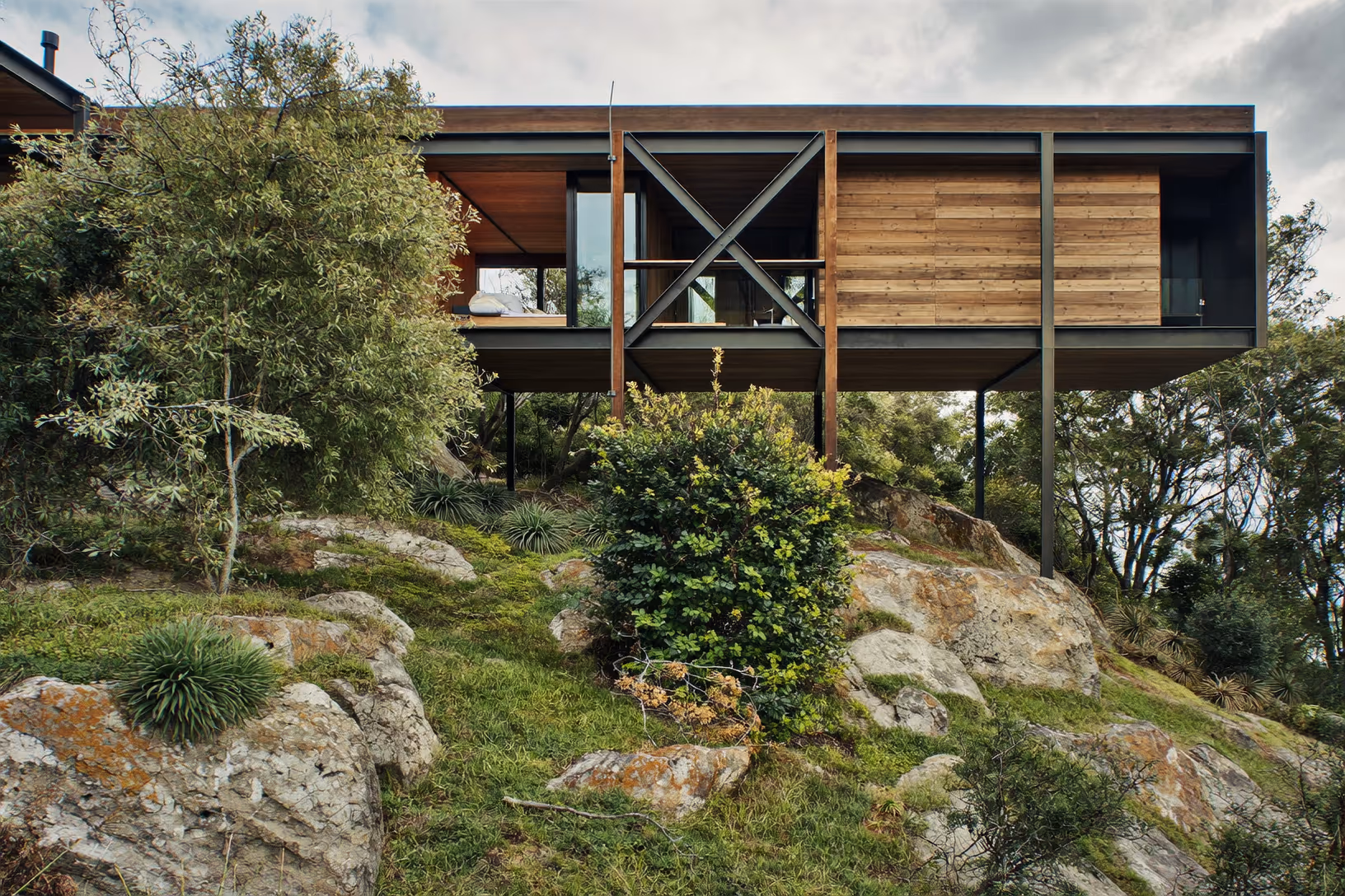 Maison sur pilotis métalliques noirs avec bardage en bois horizontal, structure en croix en acier apparent, au-dessus d'un terrain rocheux couvert de végétation dense, sous un ciel nuageux.