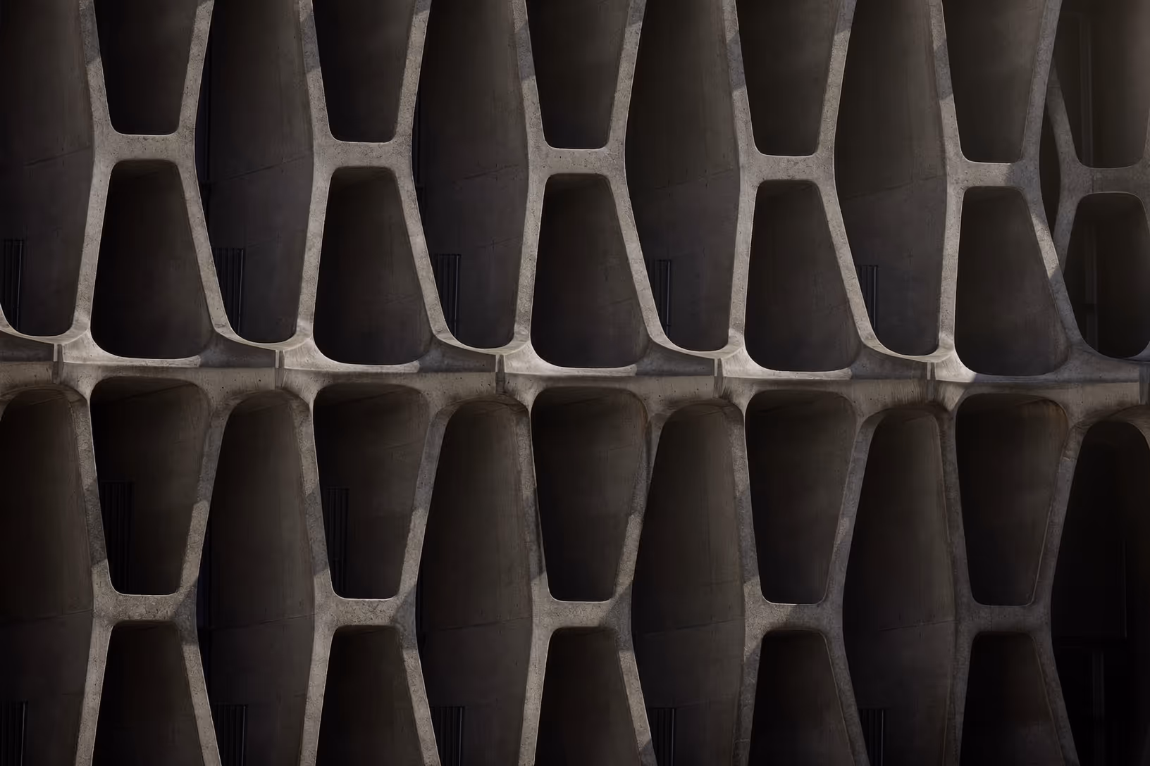 Façade en béton brut composée d'alvéoles trapézoïdales arrondies disposées en rangées superposées, photographiée en lumière rasante qui creuse des ombres profondes dans chaque vide.