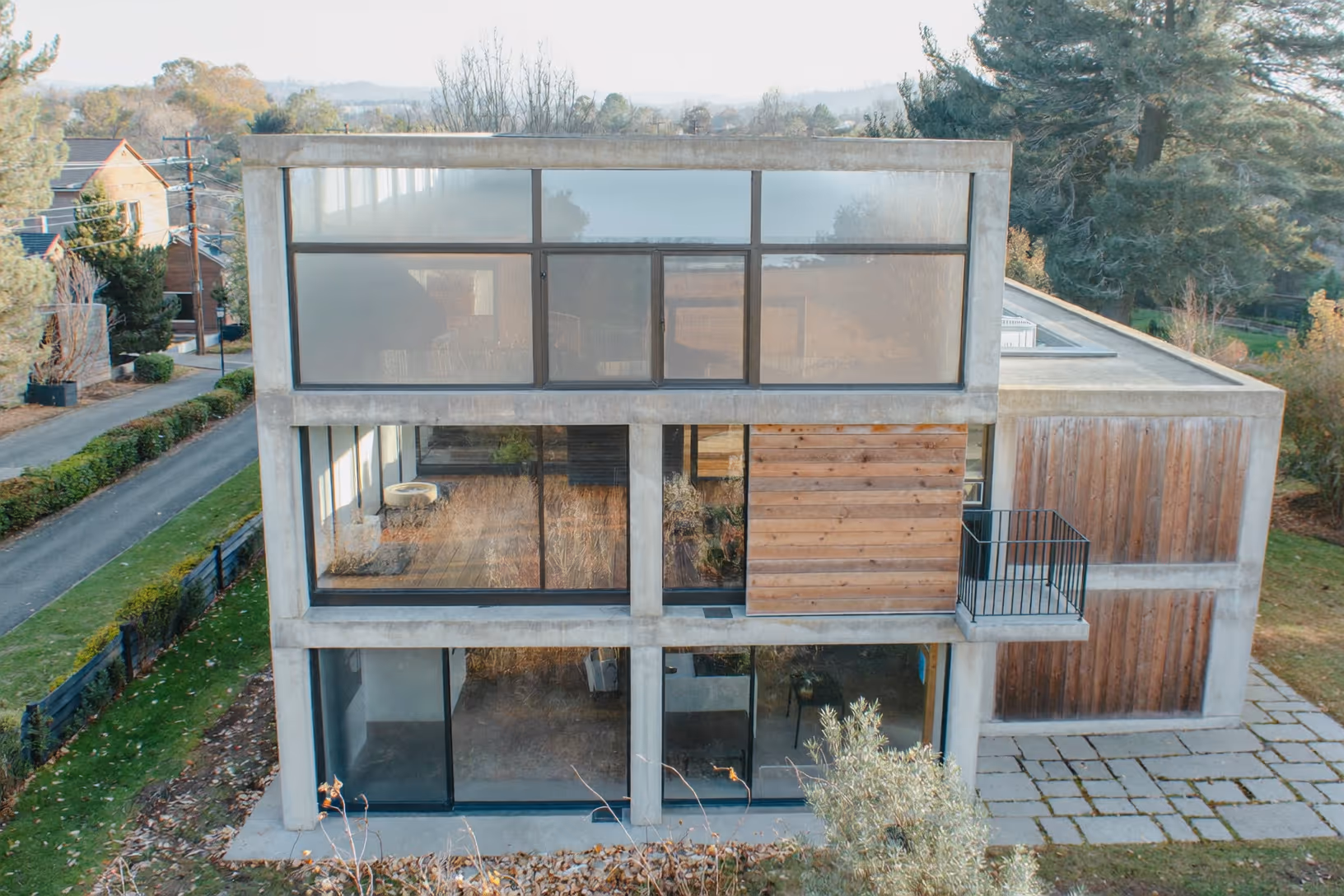 Maison de trois niveaux à structure de béton apparent, façade vitrée sur toute la hauteur, bardage bois horizontal au niveau intermédiaire, végétation automnale en arrière-plan.