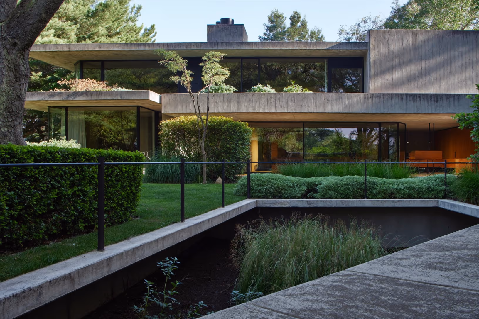 Maison à toits-dalles horizontaux en béton brut avec larges baies vitrées à cadres métalliques sombres, entourée d'un jardin structuré avec bassin rectangulaire en béton, haies taillées et grands arbres.