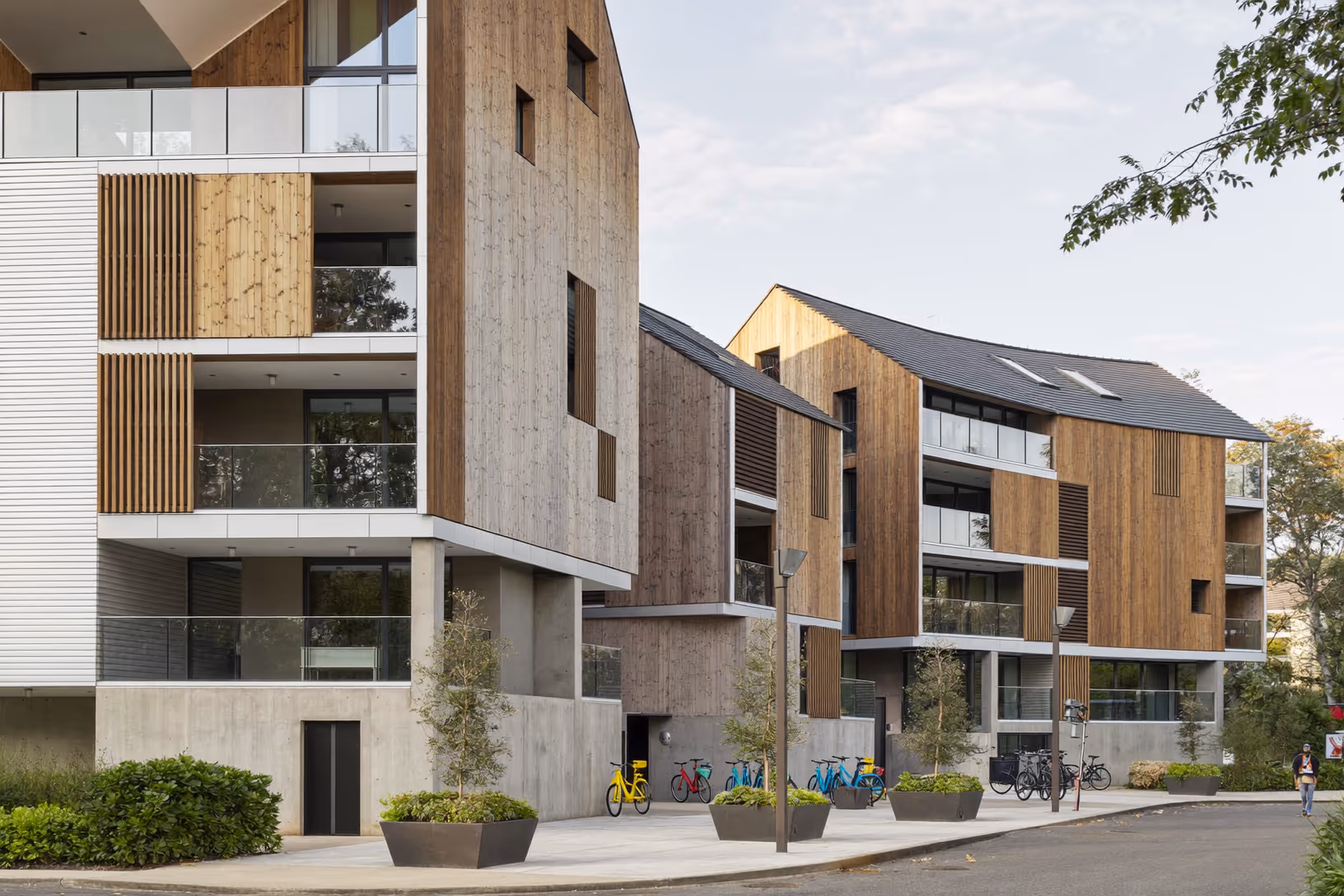 Ensemble résidentiel de plusieurs bâtiments associant béton brut en soubassement et bardage bois vertical en façade, avec toitures à deux pans sur les corps bas, des bacs plantés au pied des immeubles et des vélos garés en rez-de-chaussée.