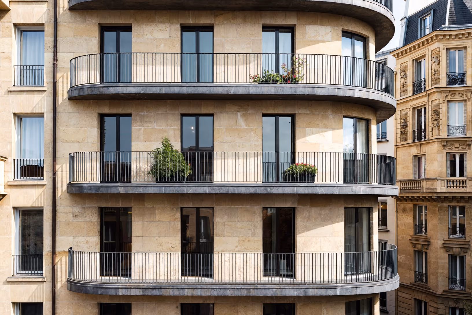 Façade contemporaine en pierre calcaire beige à balcons arrondis sur trois niveaux, garde-corps en métal noir, encadrements de fenêtres noirs et jardinières végétalisées, encadrée par deux immeubles haussmanniens.