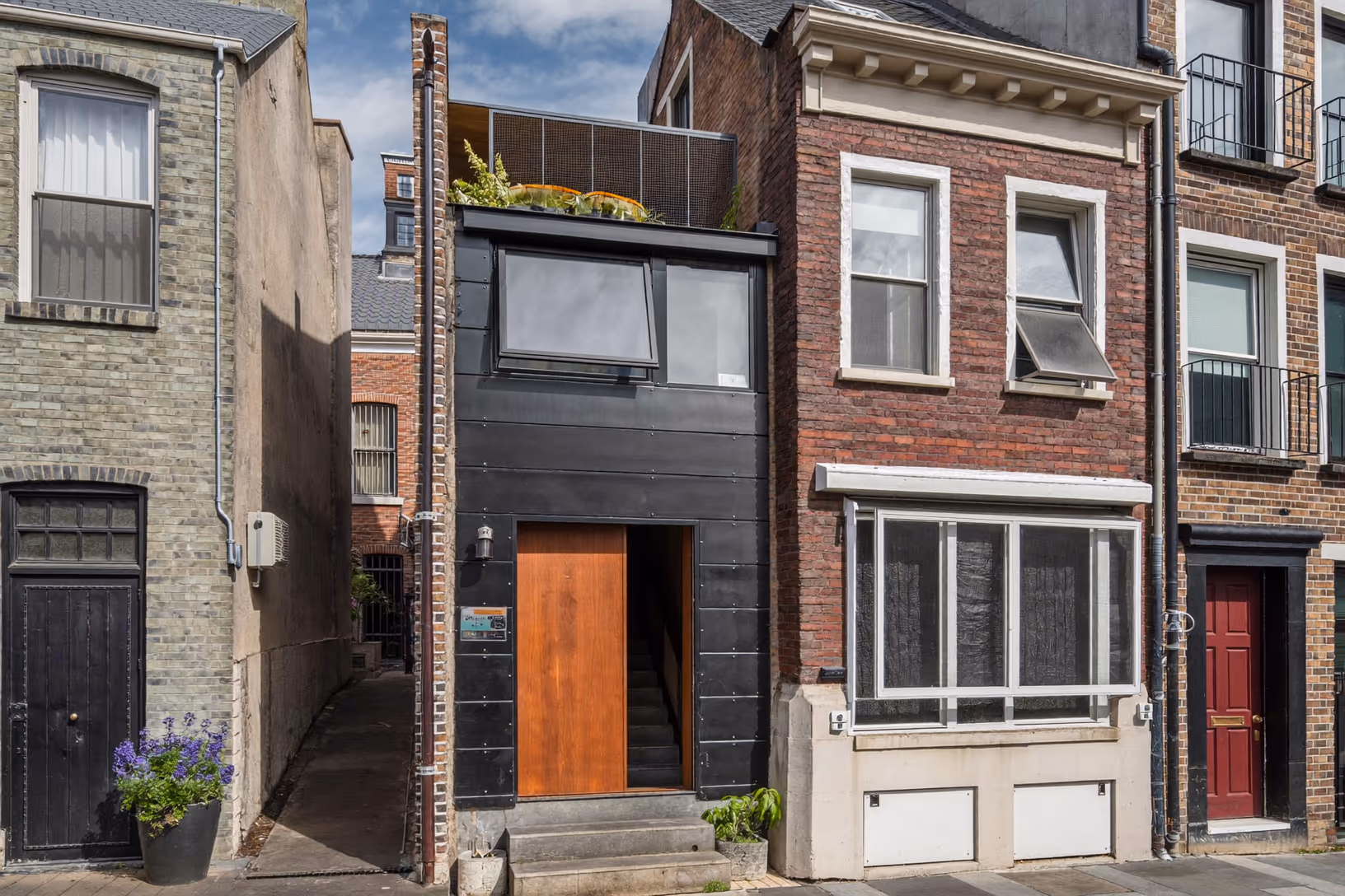 Façade étroite en métal noir mat entre deux immeubles en brique rouge, avec une porte en bois chaleureux au rez-de-chaussée et une terrasse végétalisée en toiture.