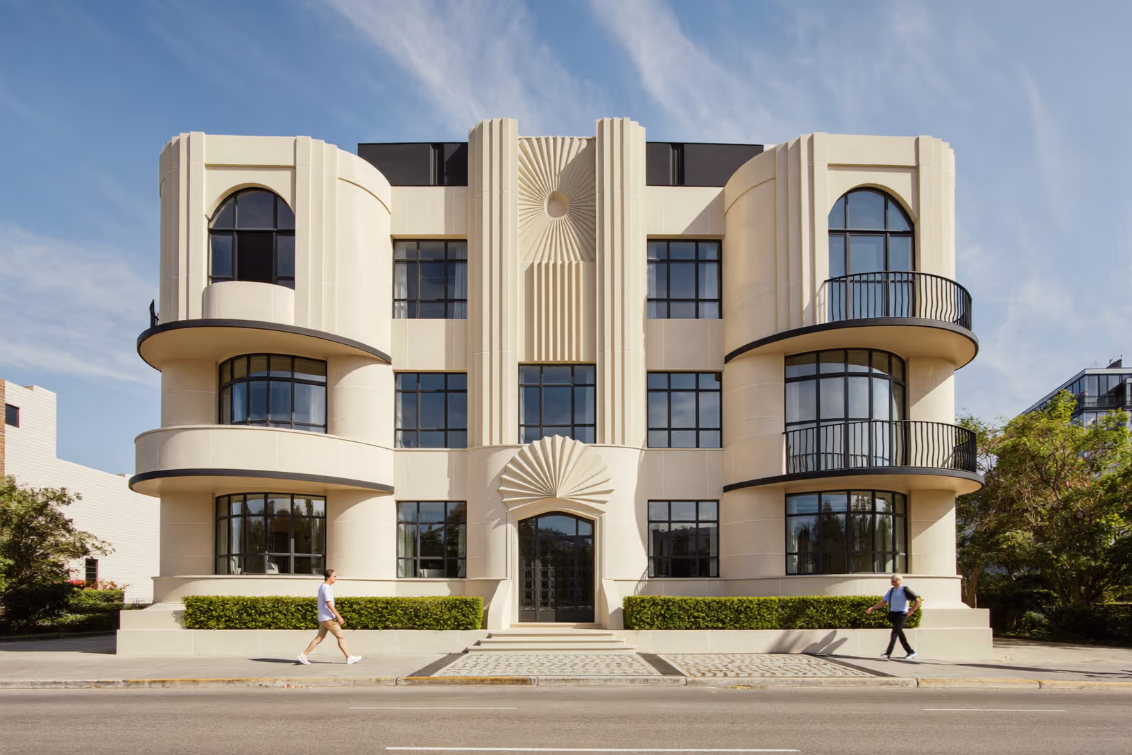 Immeuble résidentiel de trois étages à façade symétrique enduite de crème, volumes cylindriques en saillie, fenêtres arquées à menuiseries noires, décors en éventail rayonnant autour de l'entrée, haies taillées au premier plan, ciel bleu dégagé.