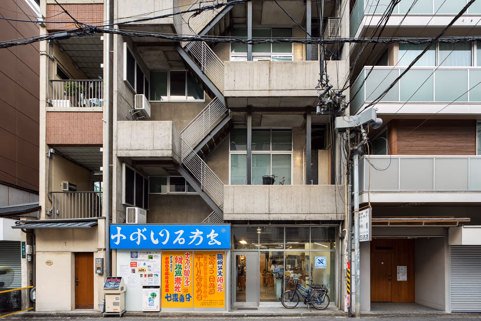 Façade frontale d'un immeuble urbain en béton brut de quatre étages, avec escalier extérieur en zigzag, balcons filants à garde-corps métallique, menuiseries aluminium, et au rez-de-chaussée une boutique aux enseignes colorées en caractères japonais. Câbles électriques en réseau dense au premier plan, ciel couvert.
